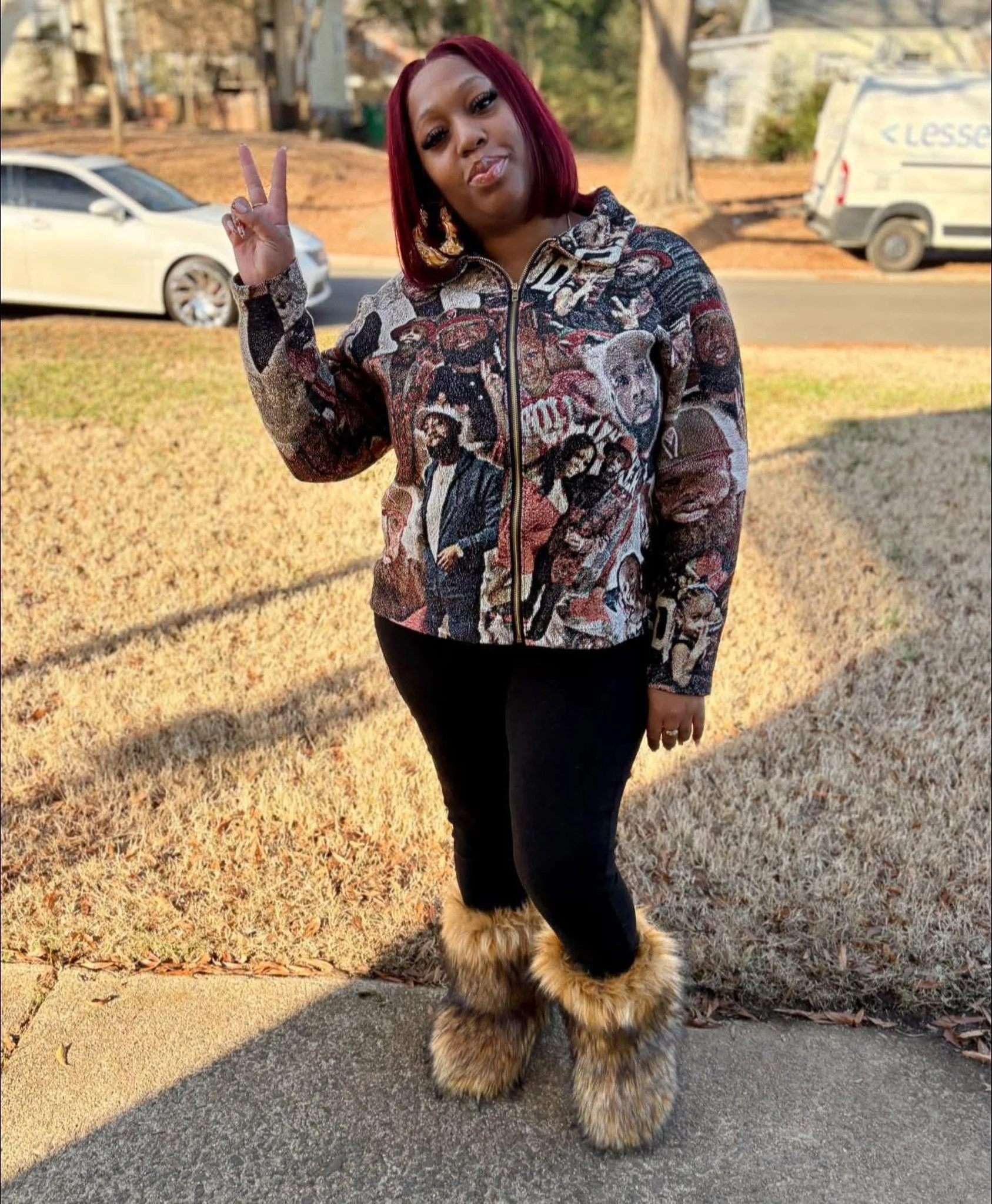 Woman with red hair wearing a sweater with art portraits and Teddy Roosevelt's face, black pants, and furry boots, standing outdoors on a sidewalk with cars and trees in the background, making a peace sign with her right hand.