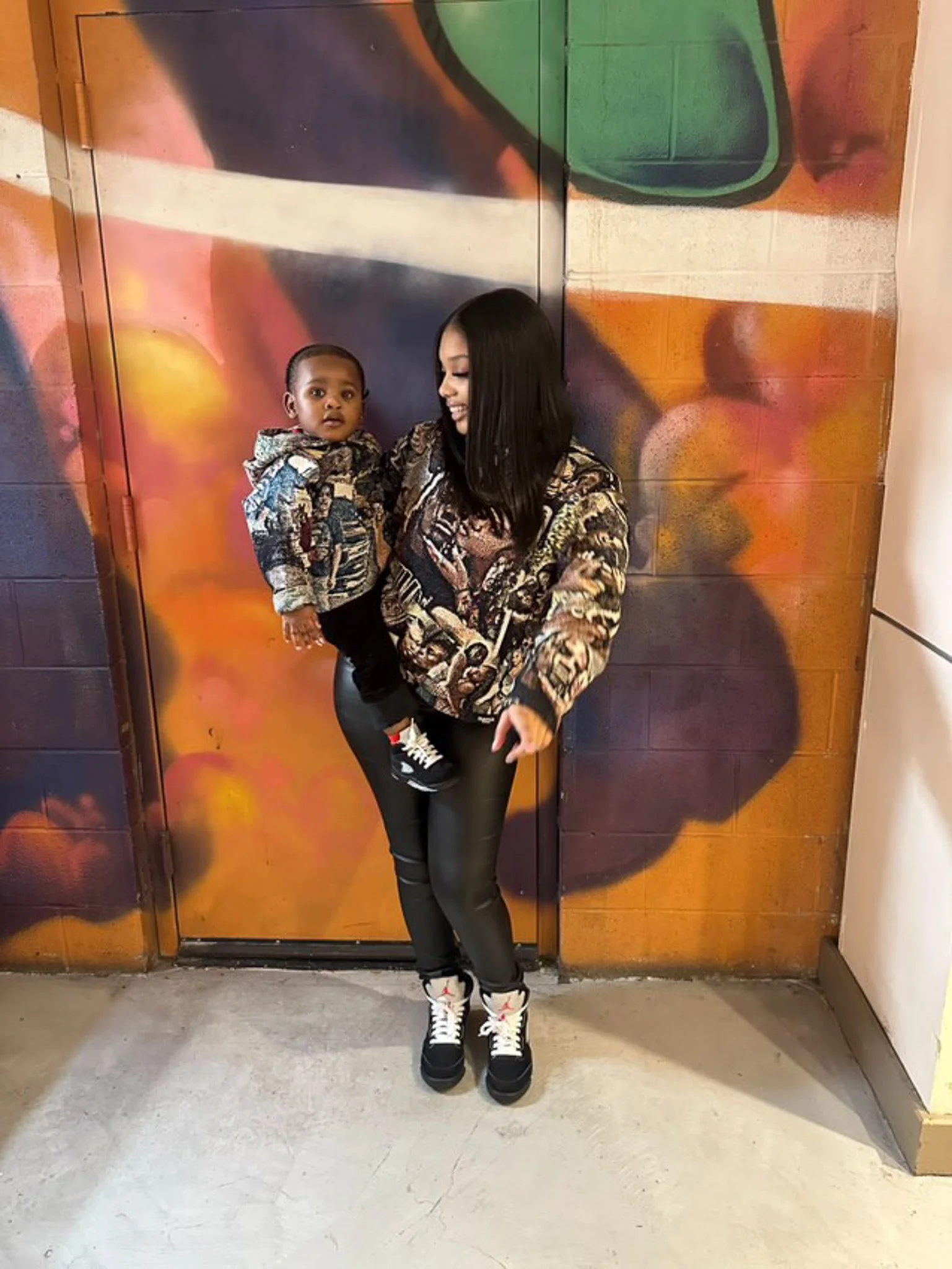 A woman holding a young child stands in front of a colorful, graffiti-style wall featuring abstract shapes and vibrant colors. The woman is smiling and looking at the child, who appears to be looking at the camera. The woman is dressed in a patterned