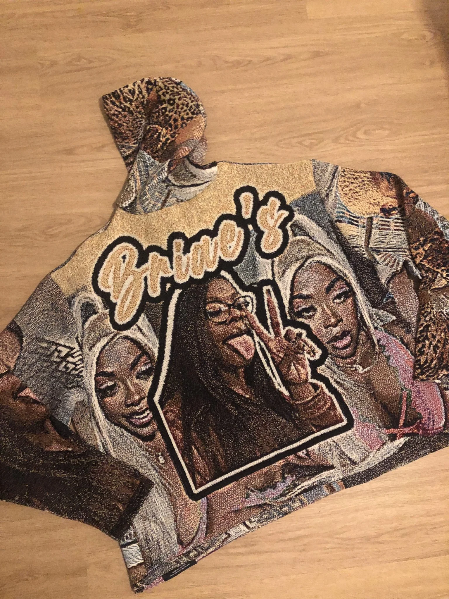 A fabric item, possibly a clothing or decorative piece, with intricate designs featuring faces, text, and patterns in shades of beige, black, and brown, laid on a wooden surface.
