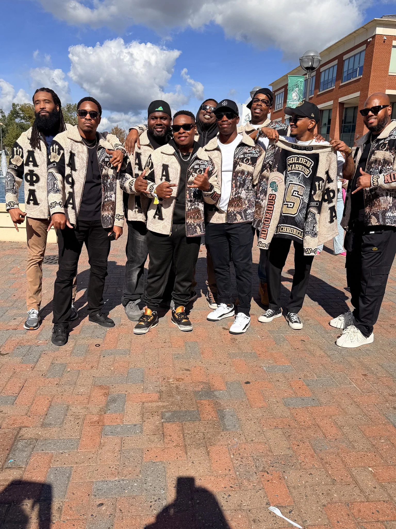 A group of nine men standing outdoors in front of a brick building, wearing matching jackets with various patches and symbols, under a partly cloudy sky.