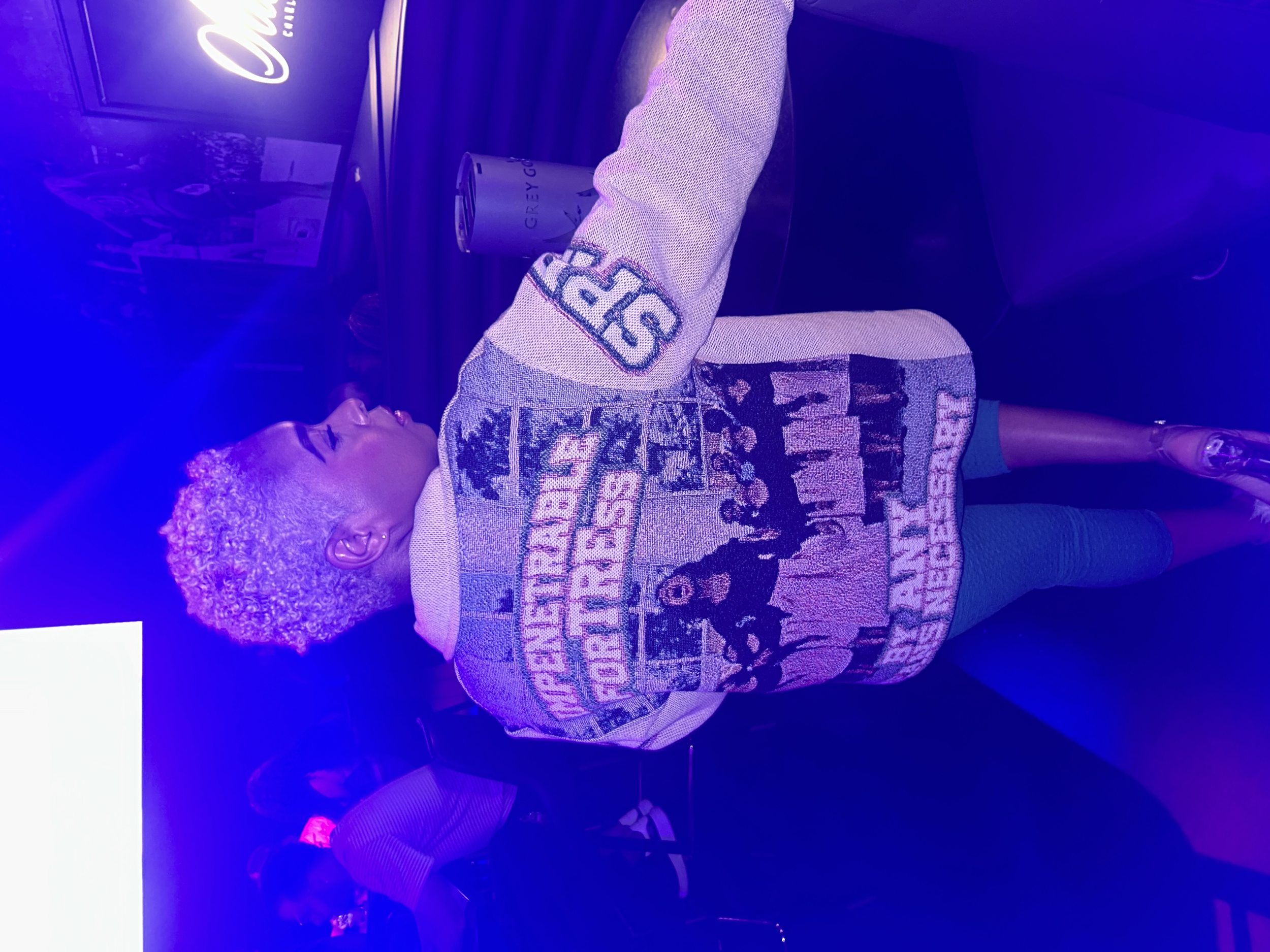 A woman with short, curly platinum blonde hair, wearing a colorful, patterned jacket and gray leggings, standing in a dimly lit space with blue and purple lighting, surrounded by other people.