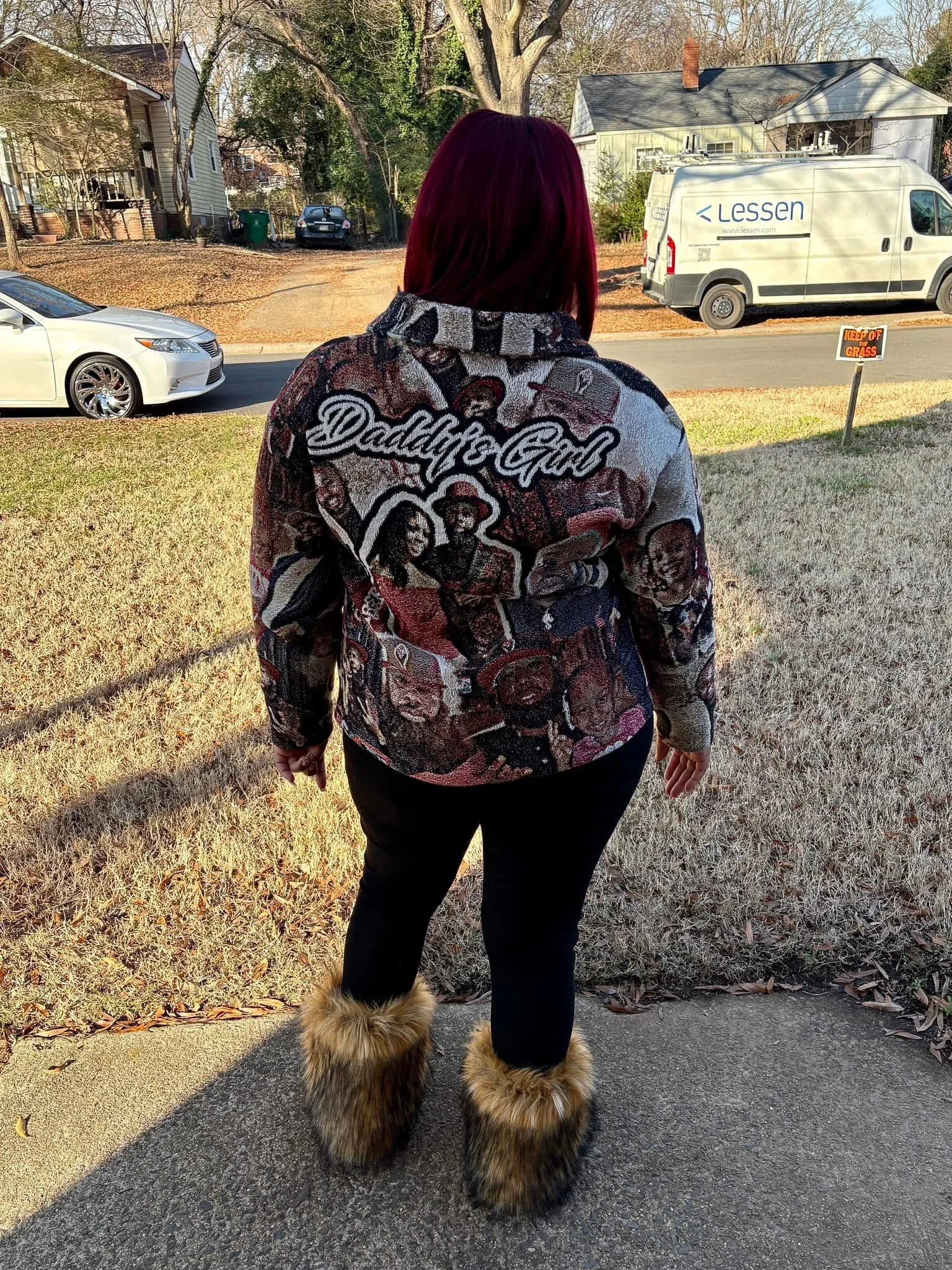 A person with long reddish hair wearing a Dundee Girl sweater, black pants, and furry shoes, standing on a sidewalk facing away from the camera. A dog with fluffy fur is visible in front of them. In the background, there is a street with cars, trees,