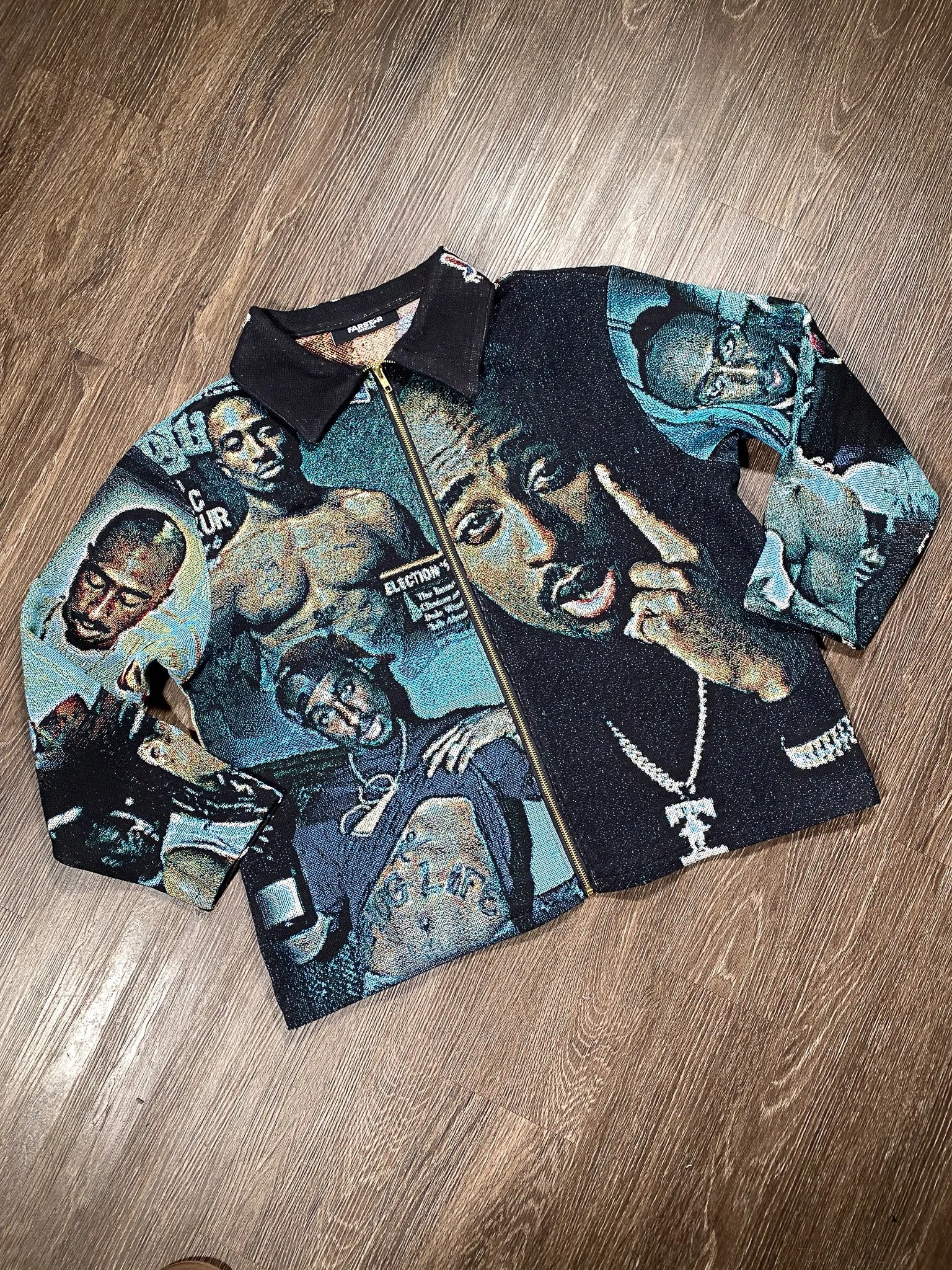 A zip-up jacket with a black collar featuring a colorful graphic design of various faces and text, laid on a wooden floor.