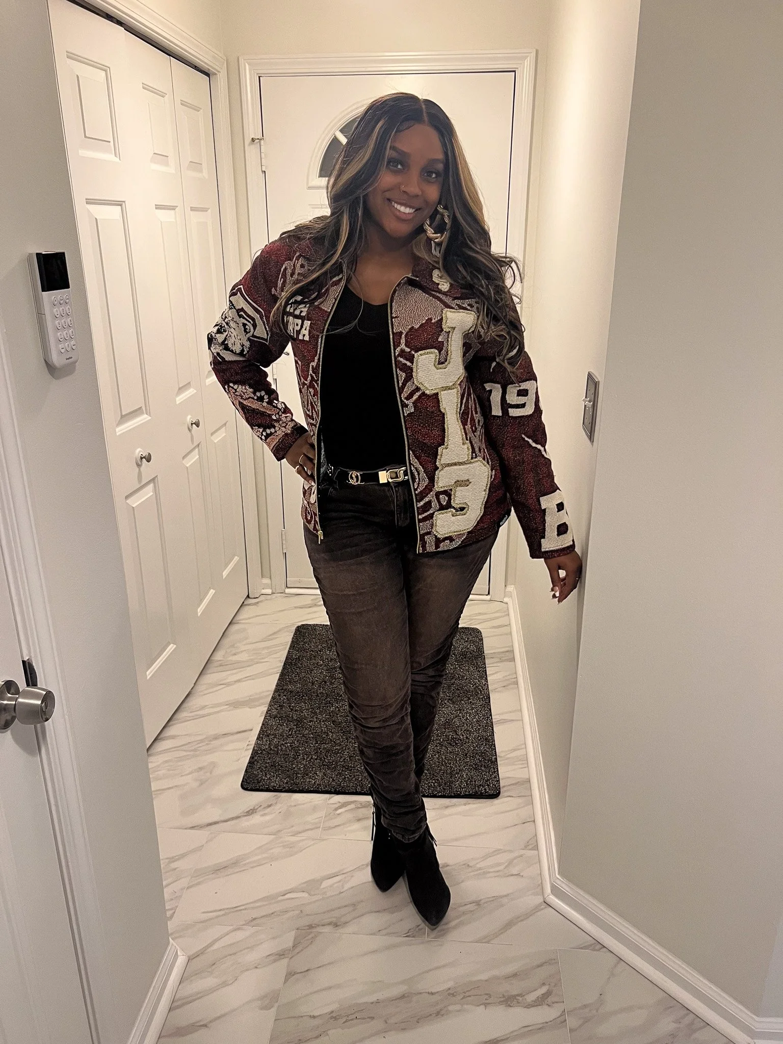 A woman with long wavy hair wearing a patchwork jacket, black top, gray pants, and black boots standing in a hallway with white walls and marble flooring, smiling at the camera.
