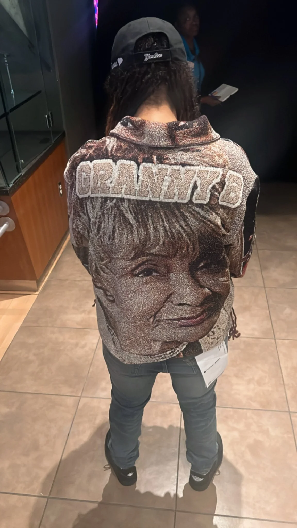 Person wearing a hoodie with a woman's face printed on the back, standing on a tiled floor inside a building.