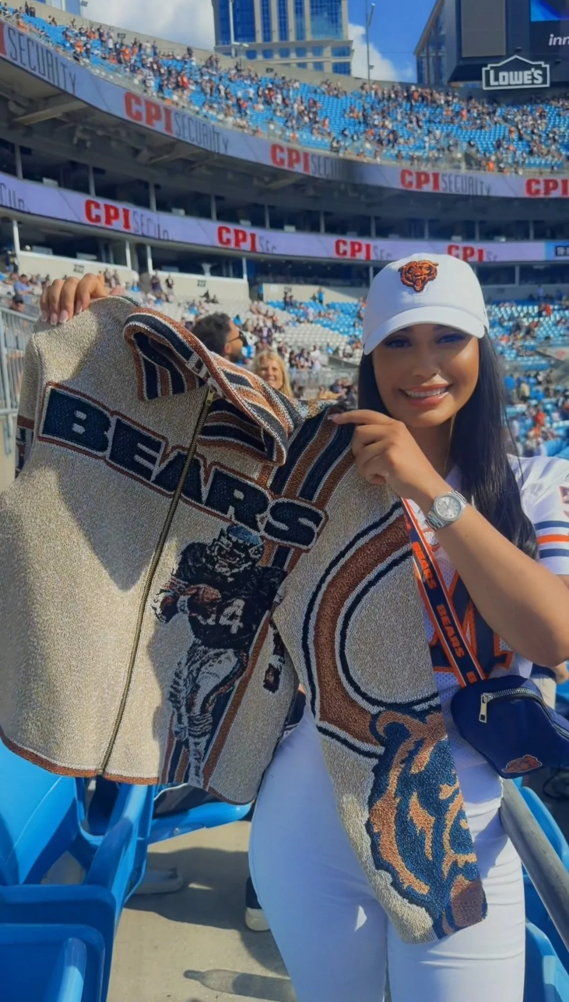 A woman smiling at a sporting event, wearing a white cap with the Chicago Bears logo, and holding a Bears scarf. She is in a stadium with blue seats and a crowd in the background.