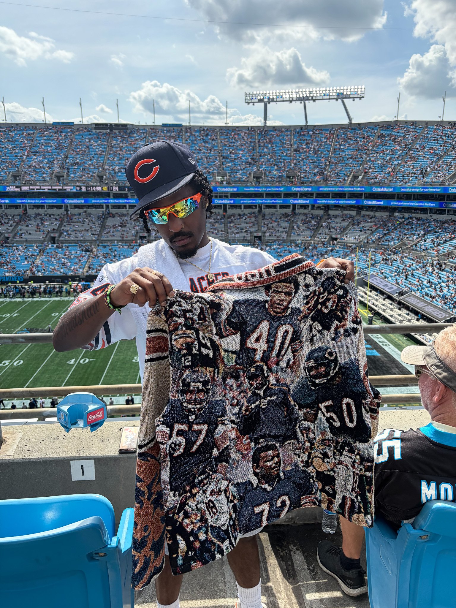 A man wearing a dark baseball cap with a red 'C' logo, reflective sunglasses, and a white sports jersey, is holding a patterned blanket with faces on it inside a stadium with blue seats and few spectators, on a partly cloudy day.