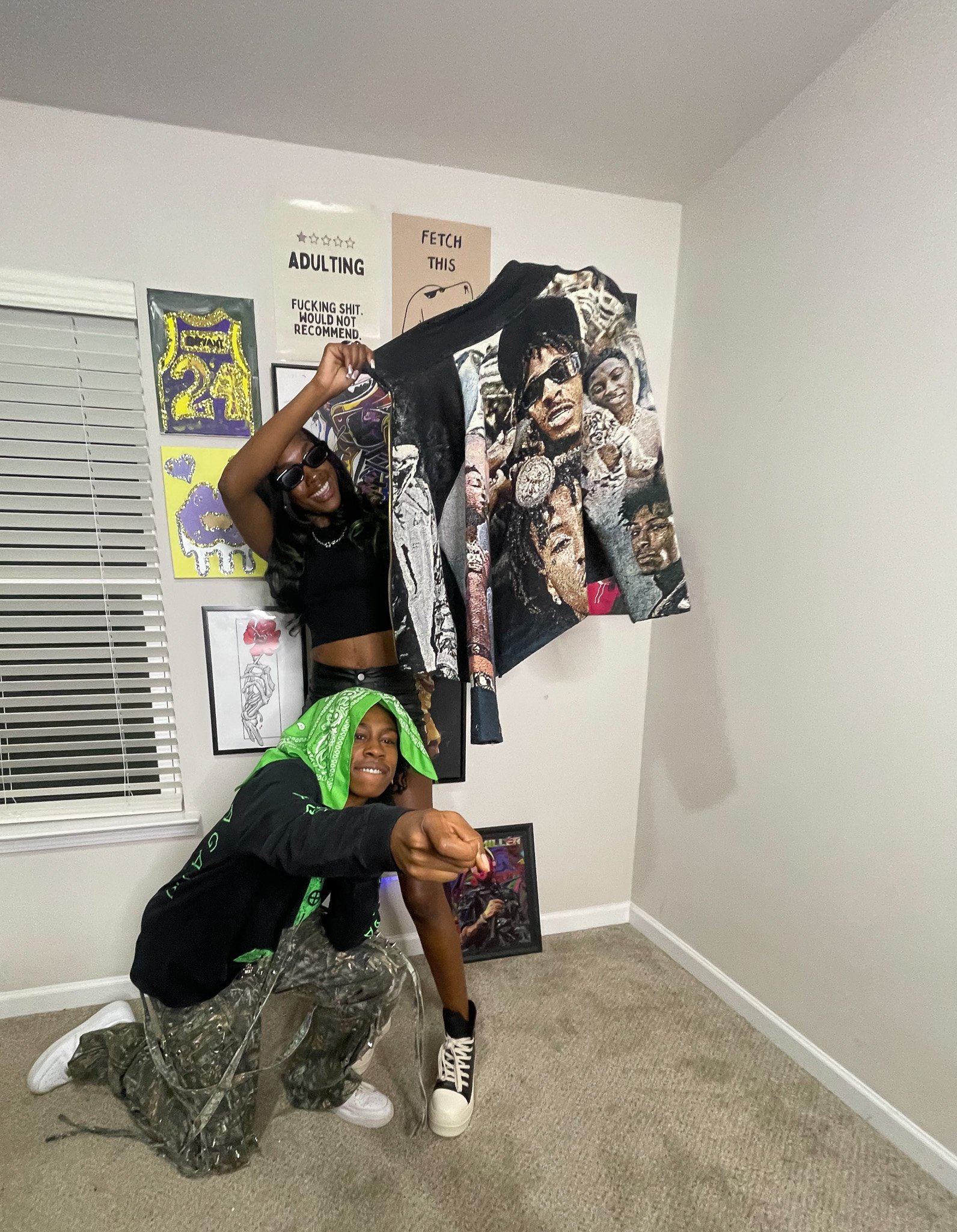 A person wearing sunglasses and a black shirt, smiling and holding an art piece, is kneeling and pointing forward. Behind them, a woman with glasses and long black hair, is standing and smiling, holding an artwork. The room has white walls decorated 