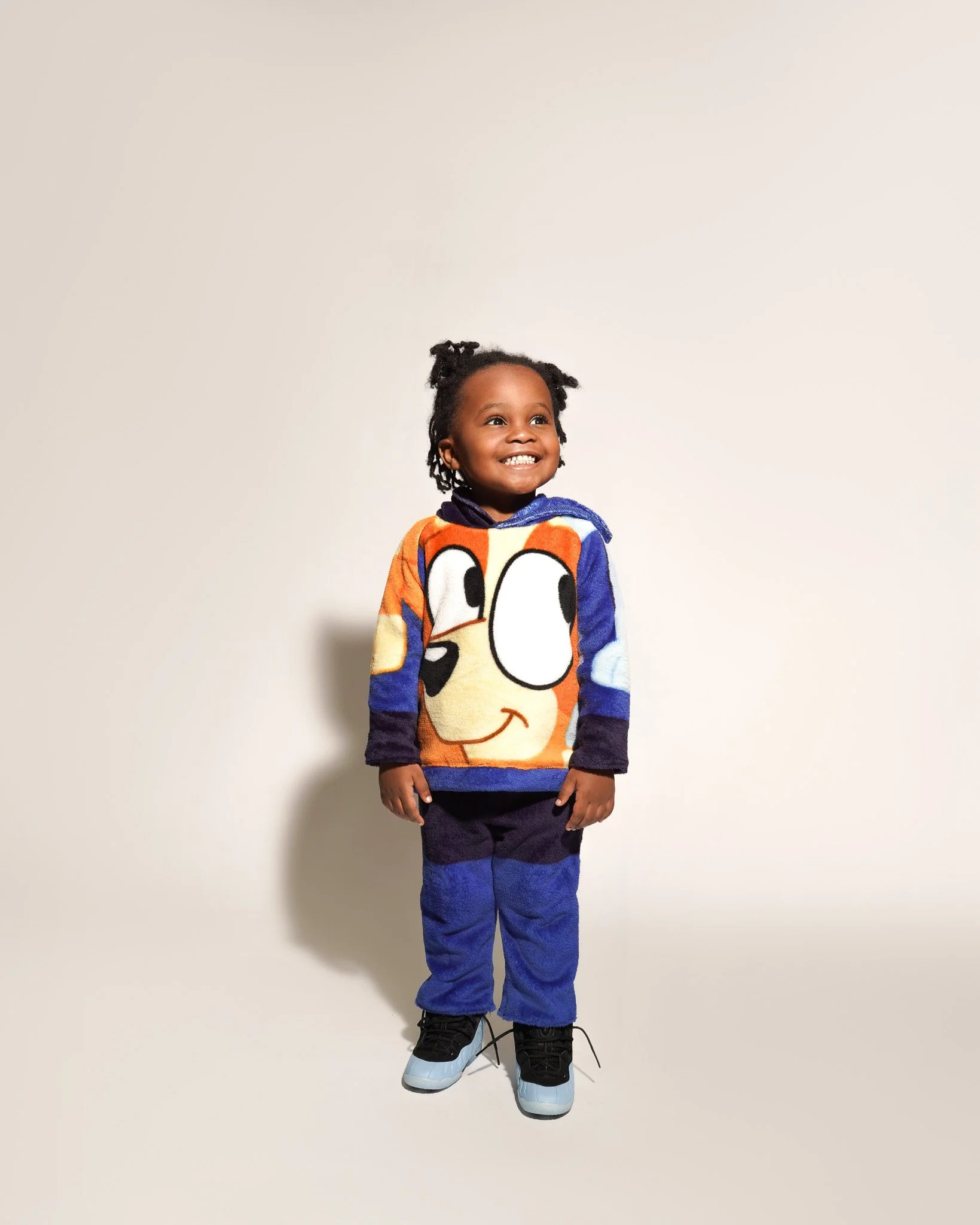 A young girl standing against a plain white background, smiling, wearing a colorful cartoon character sweater, dark pants, and sneakers.