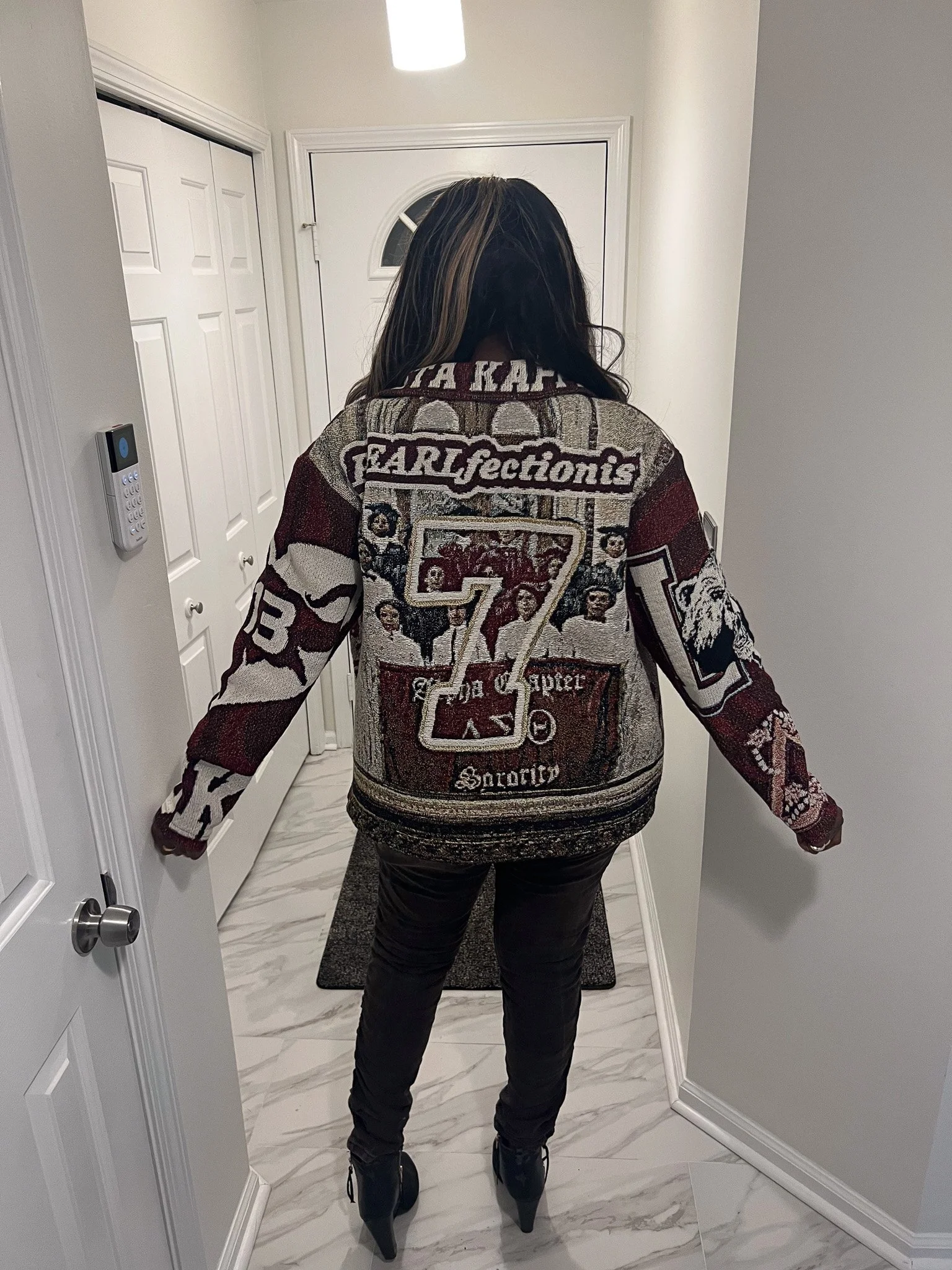 Person standing in a hallway, viewed from the back, wearing a patterned jacket and black high-heeled boots.