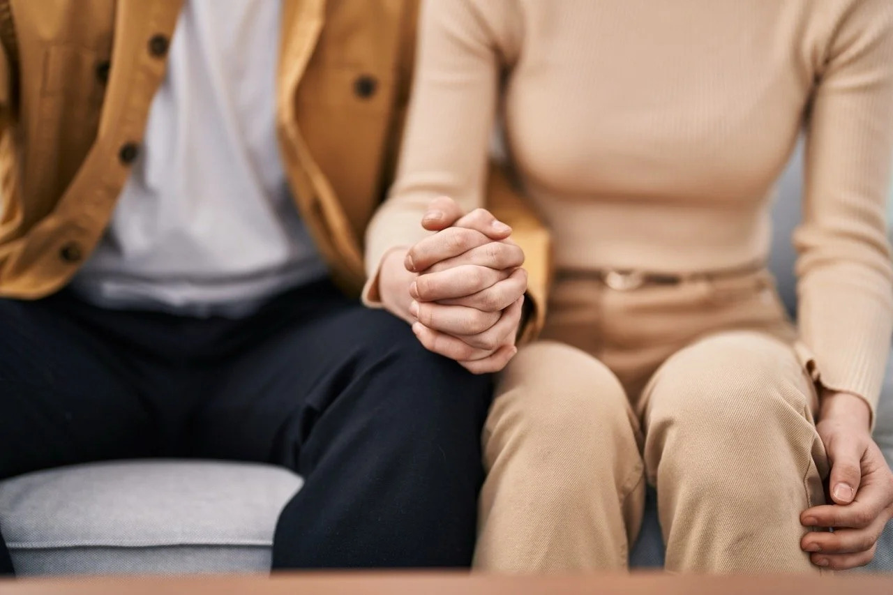 Two people sitting on a couch, holding hands with fingers intertwined.
