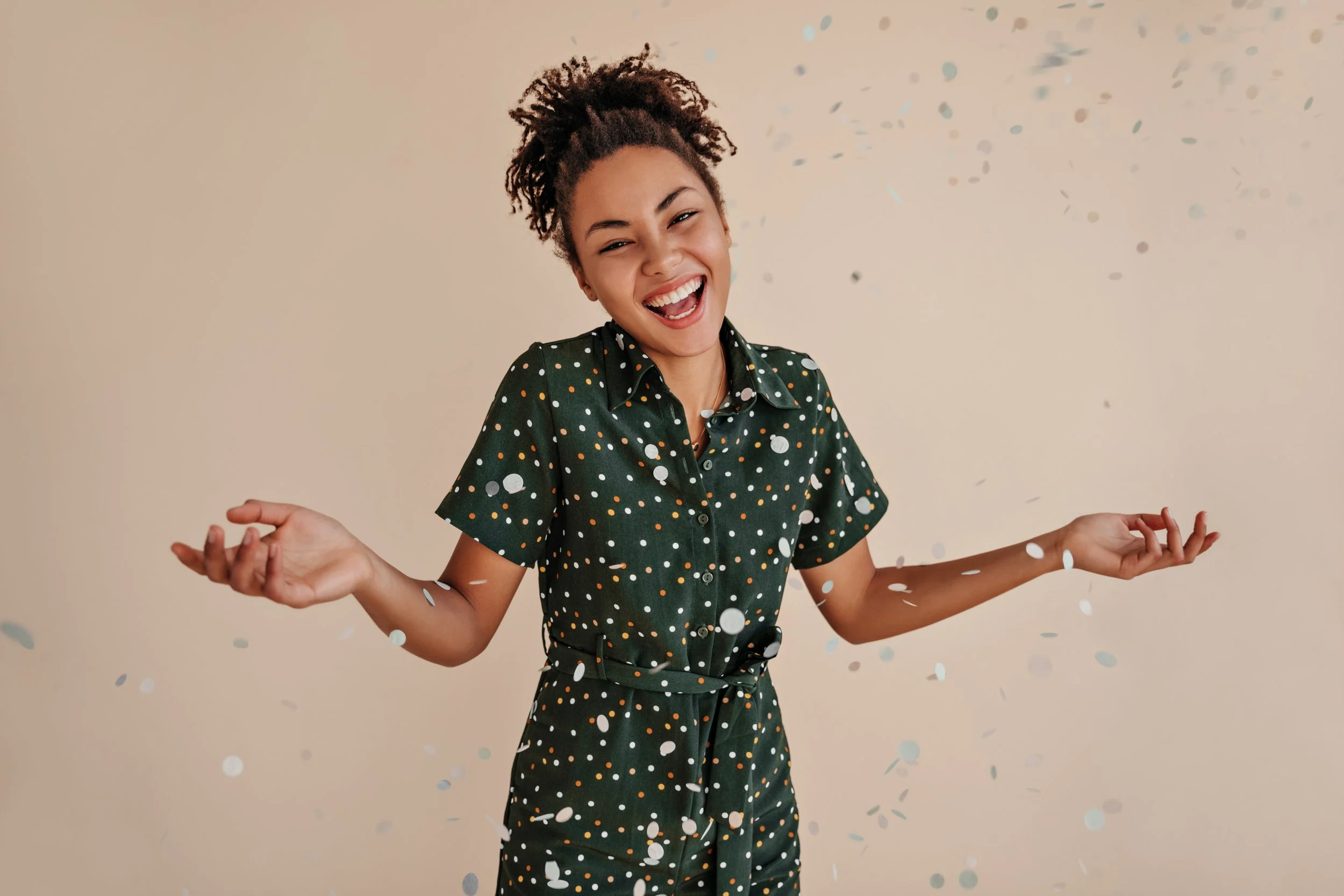 A young woman with curly hair wearing a green polka-dot jumpsuit, smiling and celebrating amidst falling confetti.