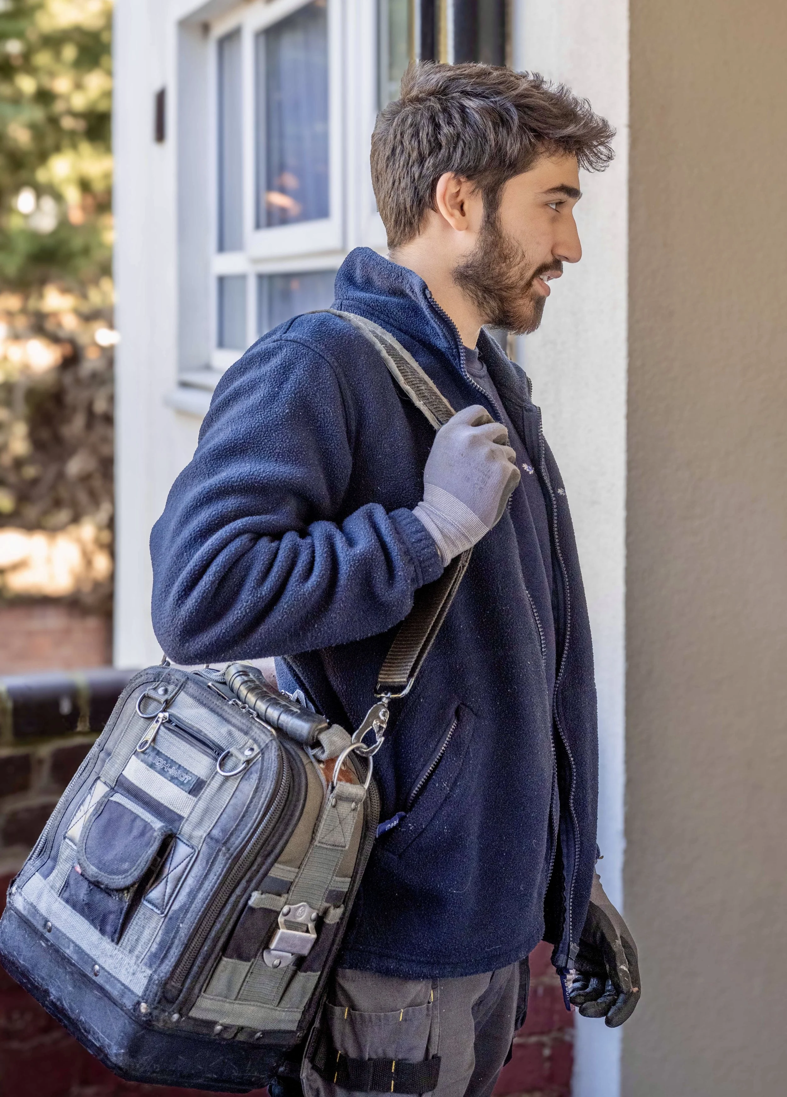 A young man with a beard, wearing a navy fleece and gloves, carrying a tool bag and a backpack, standing outside a house and looking at a wall.