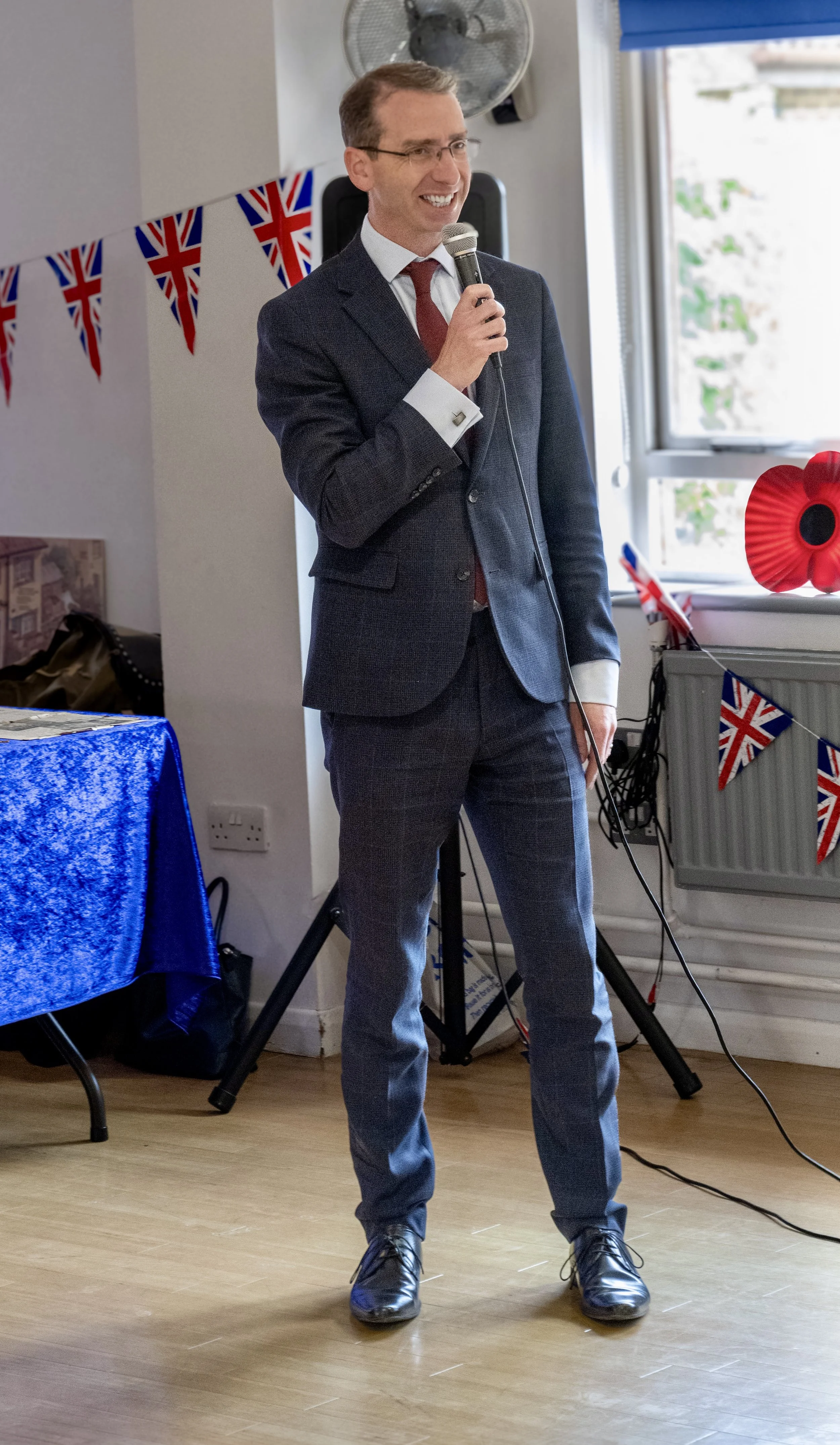 Man in a suit holding a microphone giving a speech at a decorated indoor event with UK flags and patriotic decorations.