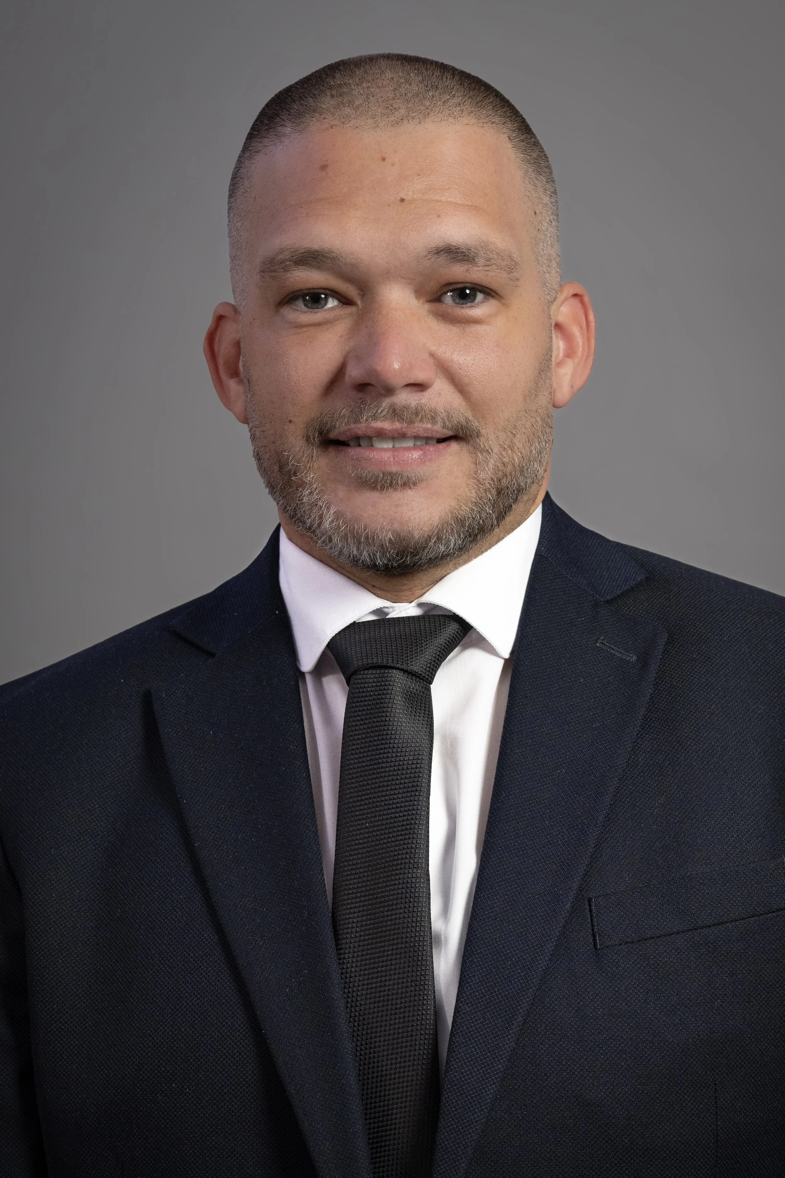 Headshot of a smiling man in a dark suit, white shirt, and black tie against a grey background.Corporate Headshot