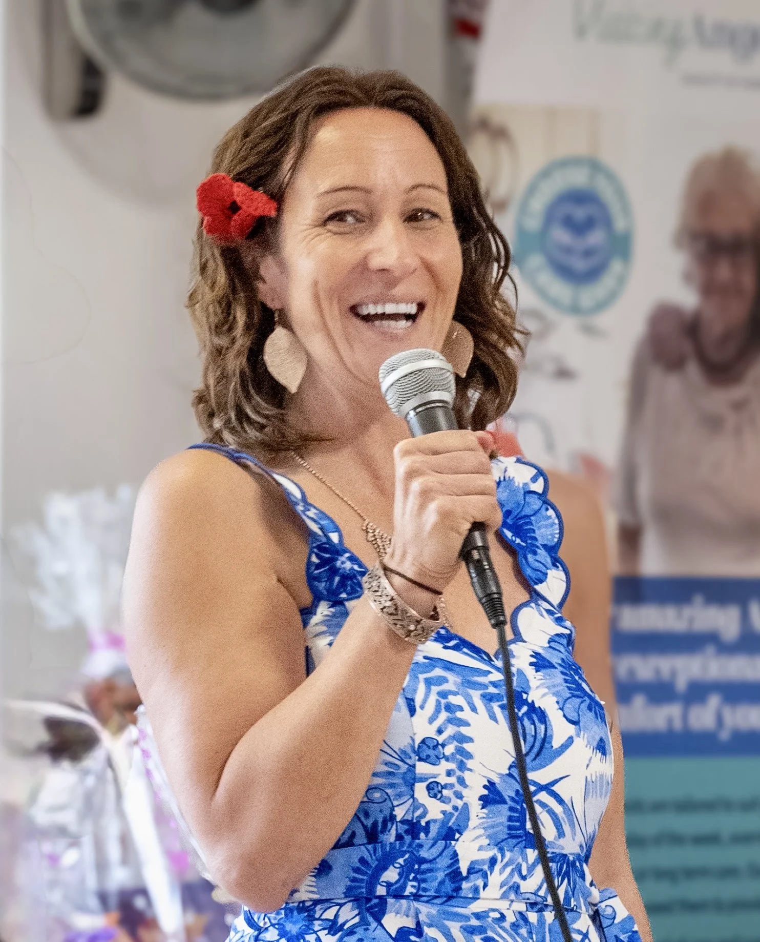 A woman with brown hair wearing a blue and white floral dress, holding a microphone, and speaking at an indoor event. She has a red flower hair accessory and large earrings, smiling and looking slightly to her right.