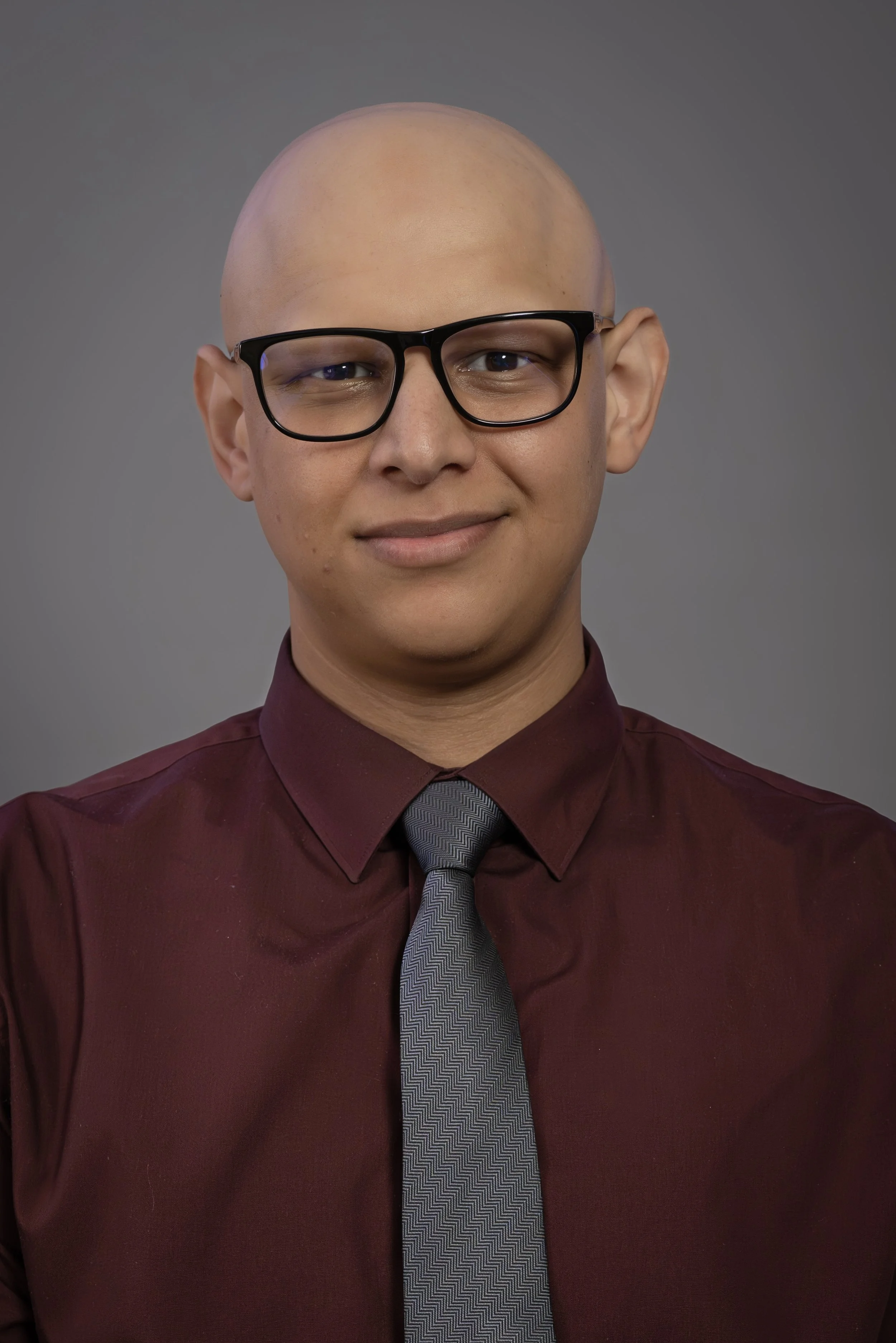 Portrait of a young man with glasses, dressed in a maroon shirt and grey tie, against a grey background.Corporate Headshot