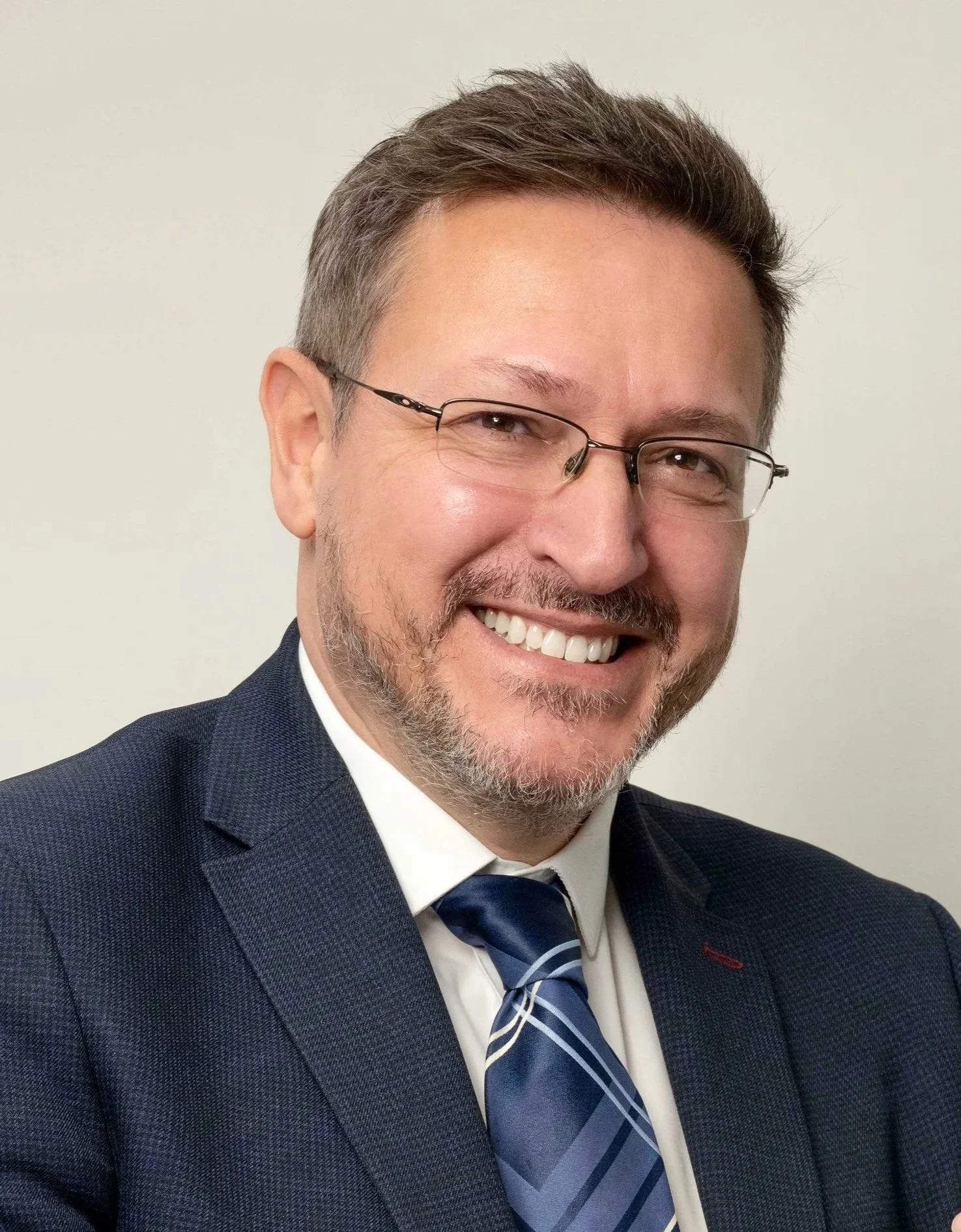 A smiling man wearing glasses, a dark blue suit, white shirt, and a blue patterned tie, adjusting his wristwatch against a plain white background.