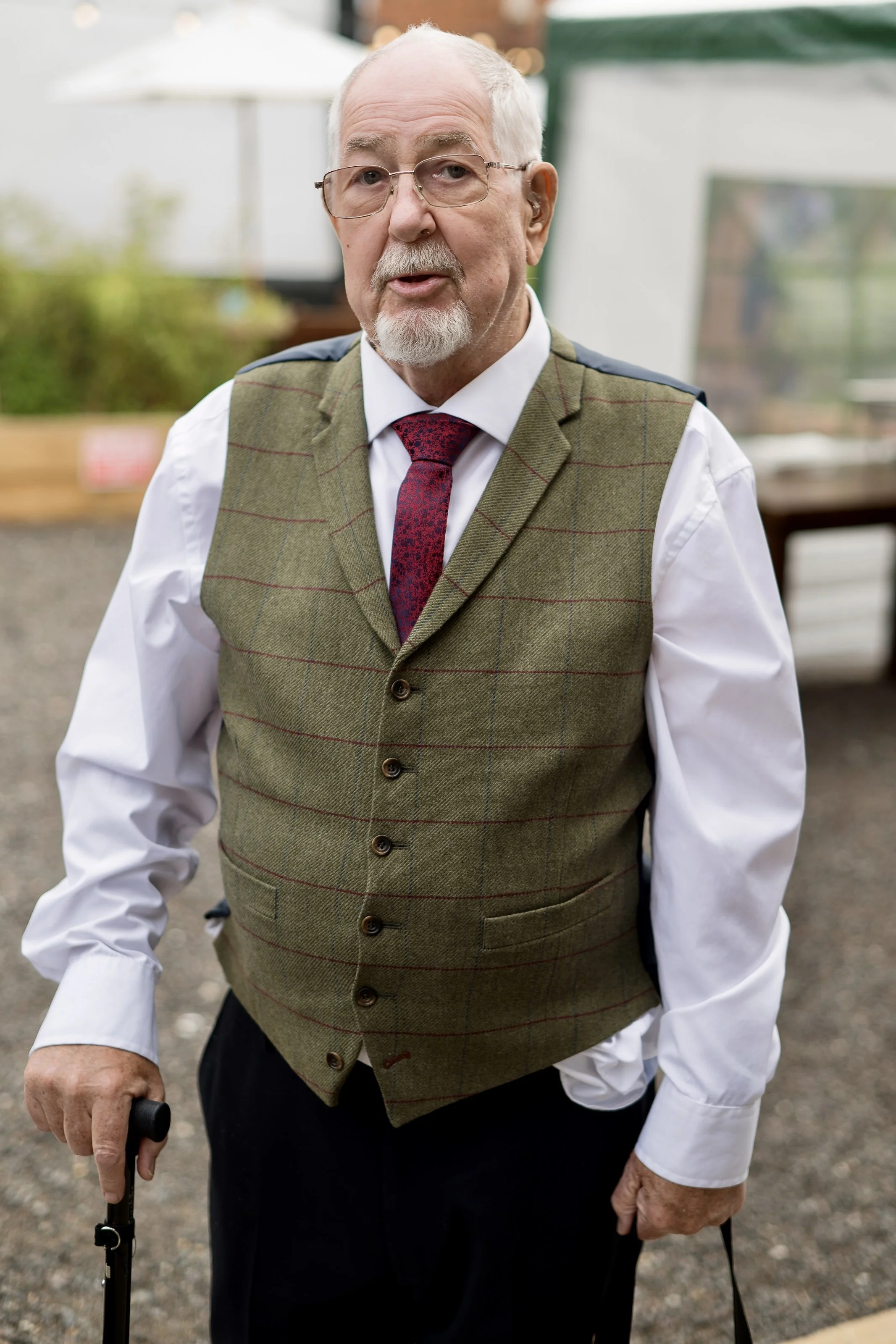 An elderly man with glasses and a beard, wearing a green plaid vest, white shirt, and red tie, standing outdoors with a cane in his right hand.