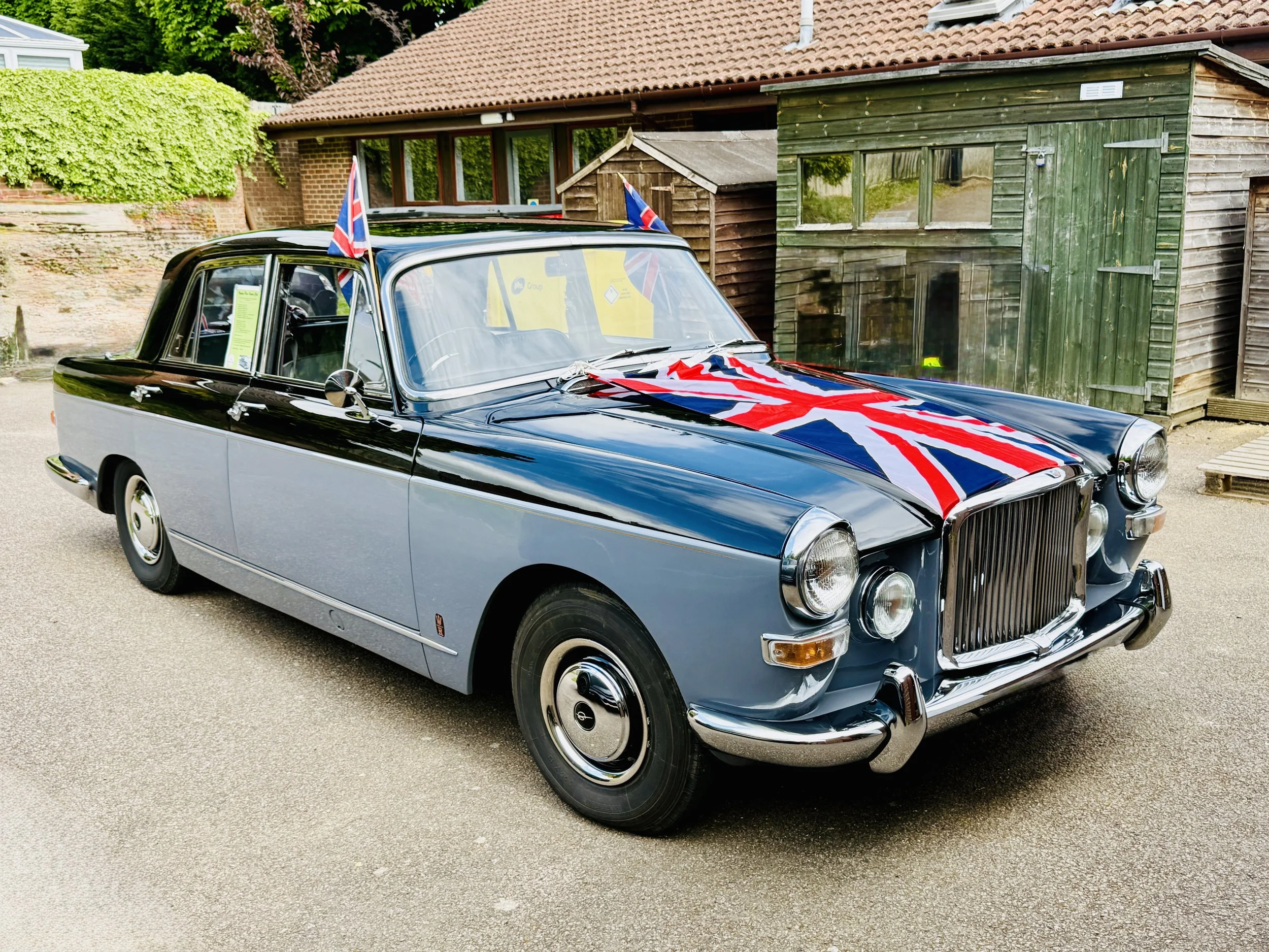 A vintage black and silver car with Union Jack flags on the roof and a large Union Jack flag draped over the hood, parked on a driveway near wooden sheds.