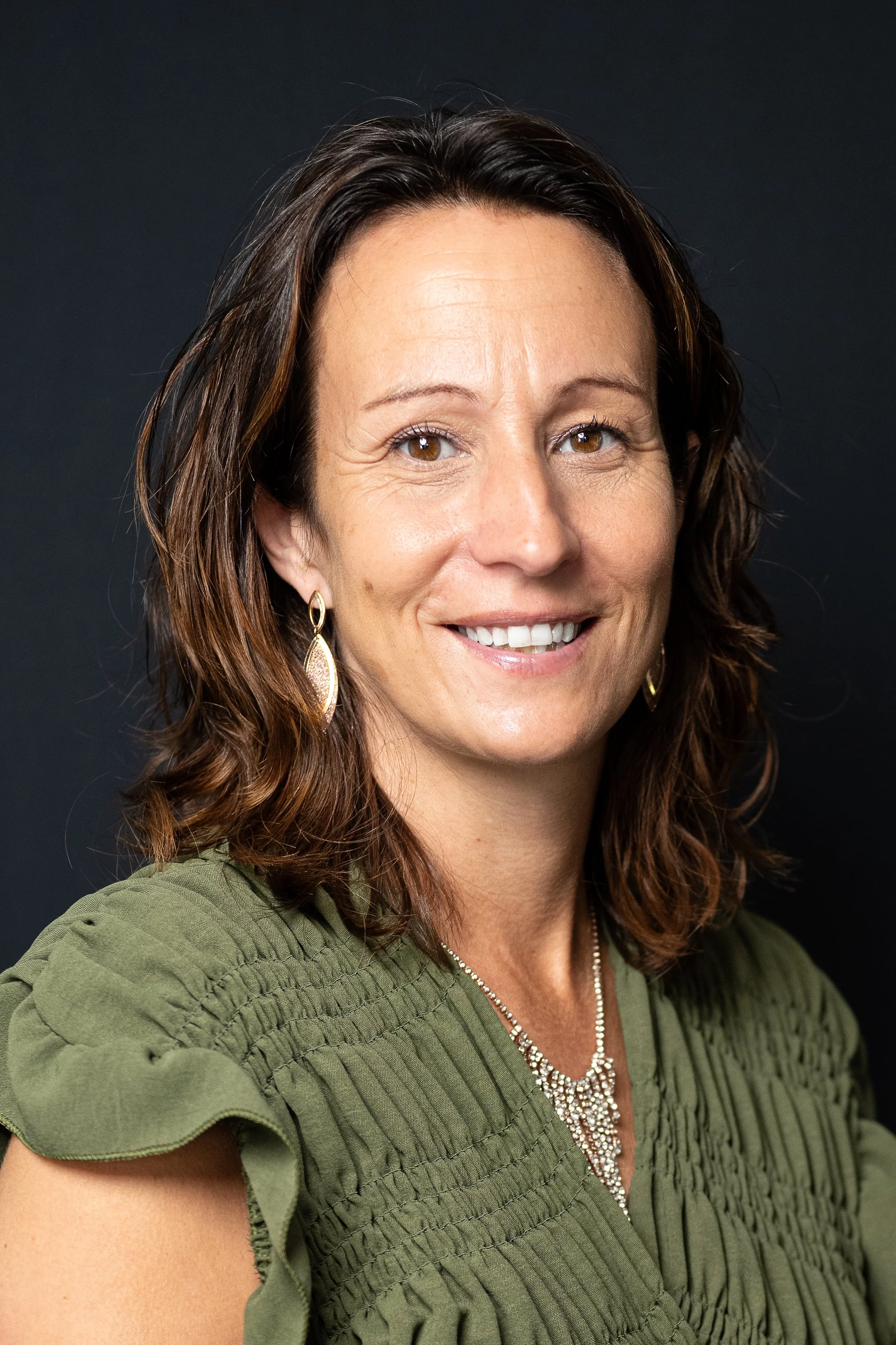 A woman with shoulder-length brown hair, light skin, and brown eyes, smiling at the camera. She is wearing gold earrings, a silver necklace, and a green blouse with gathered fabric on the shoulders, set against a dark background.