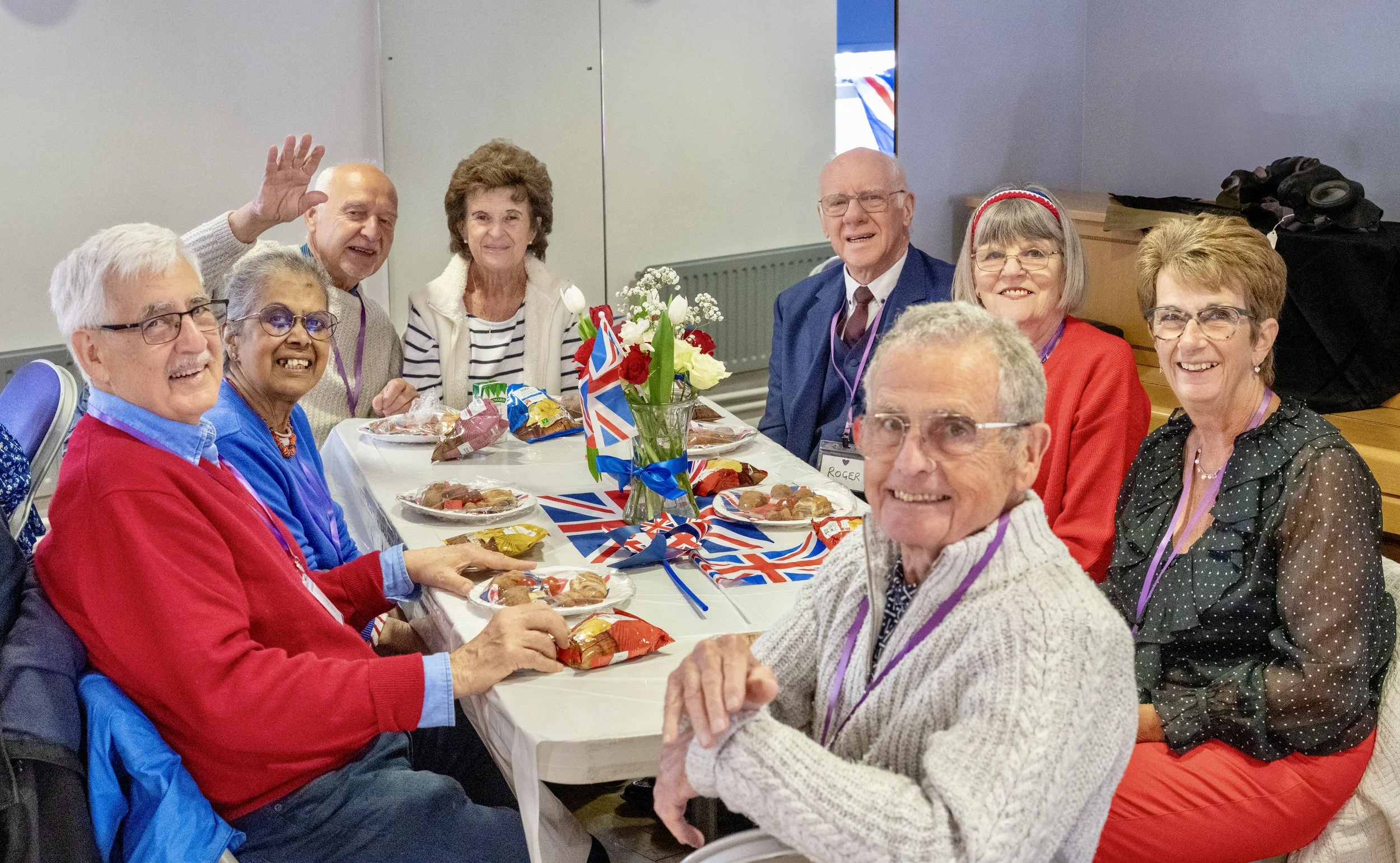 Group of elderly people sitting around a table celebrating with Union Jack flags, food, and drinks, in a room with gray walls.