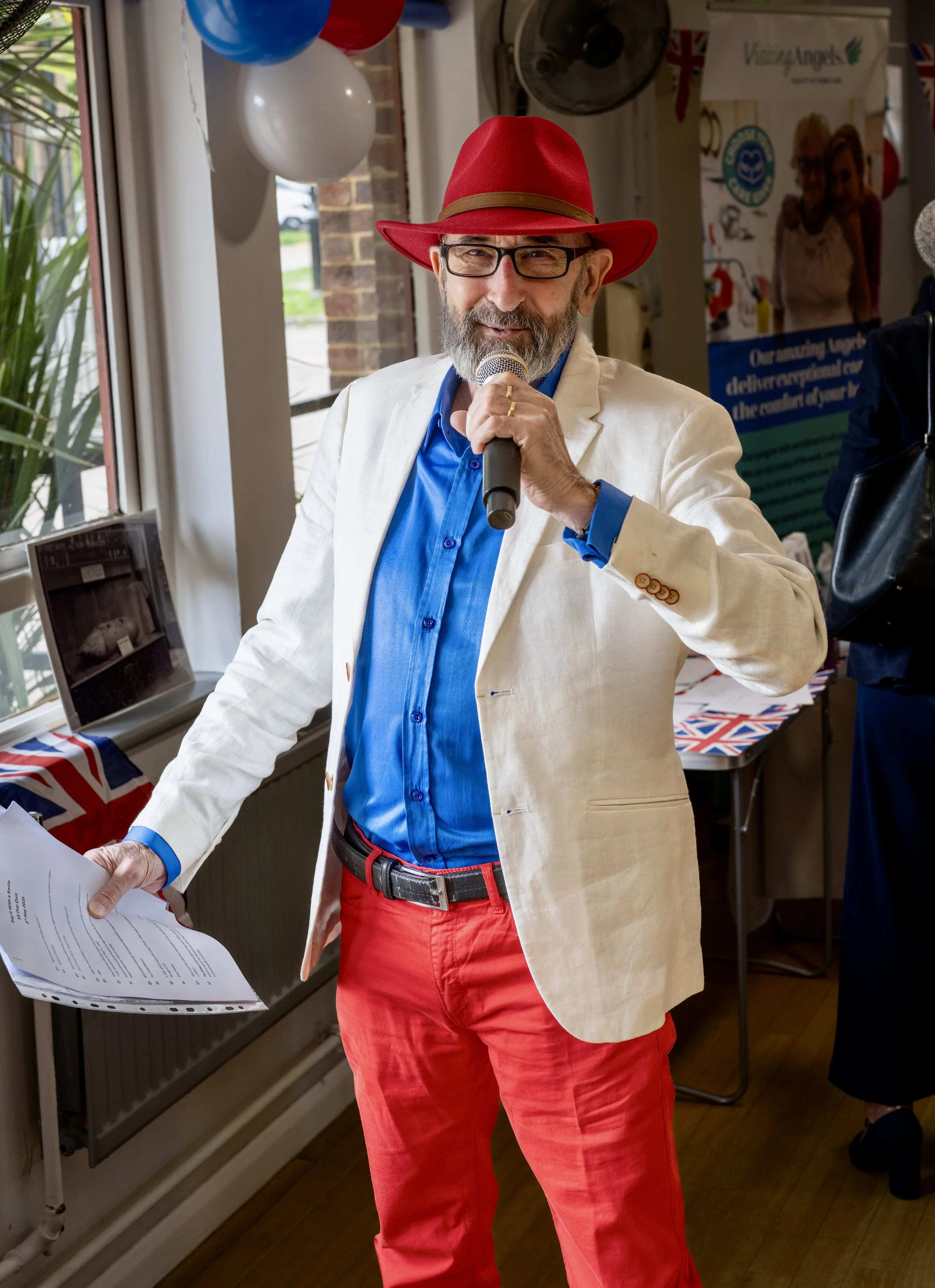 Man wearing a red hat, glasses, a white blazer, and red pants holding a microphone and papers, speaking at an indoor event."}