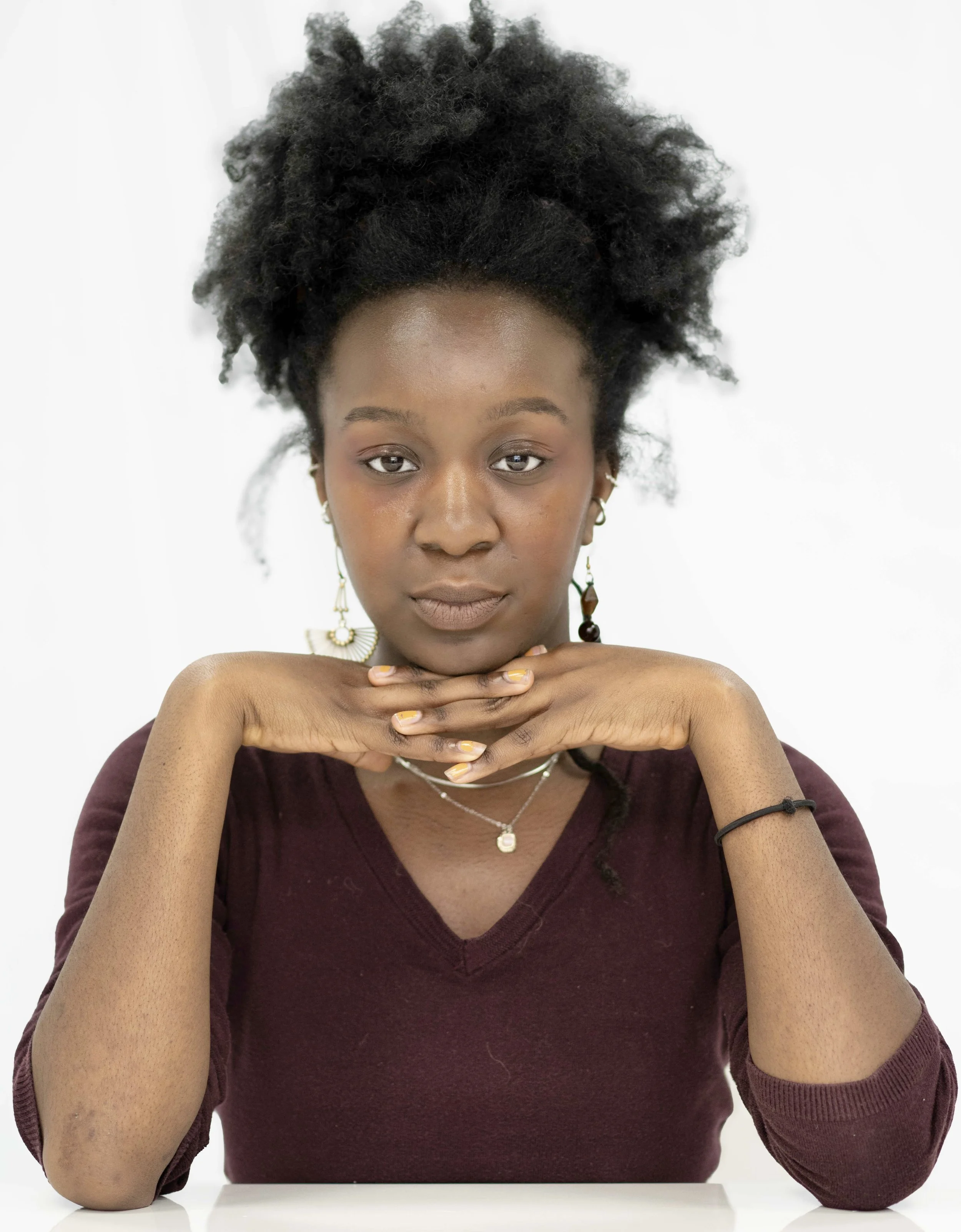 A young woman with natural curly hair, wearing a maroon long-sleeve shirt, jewelry, and sitting with her hands under her chin, looking directly at the camera against a plain white background.