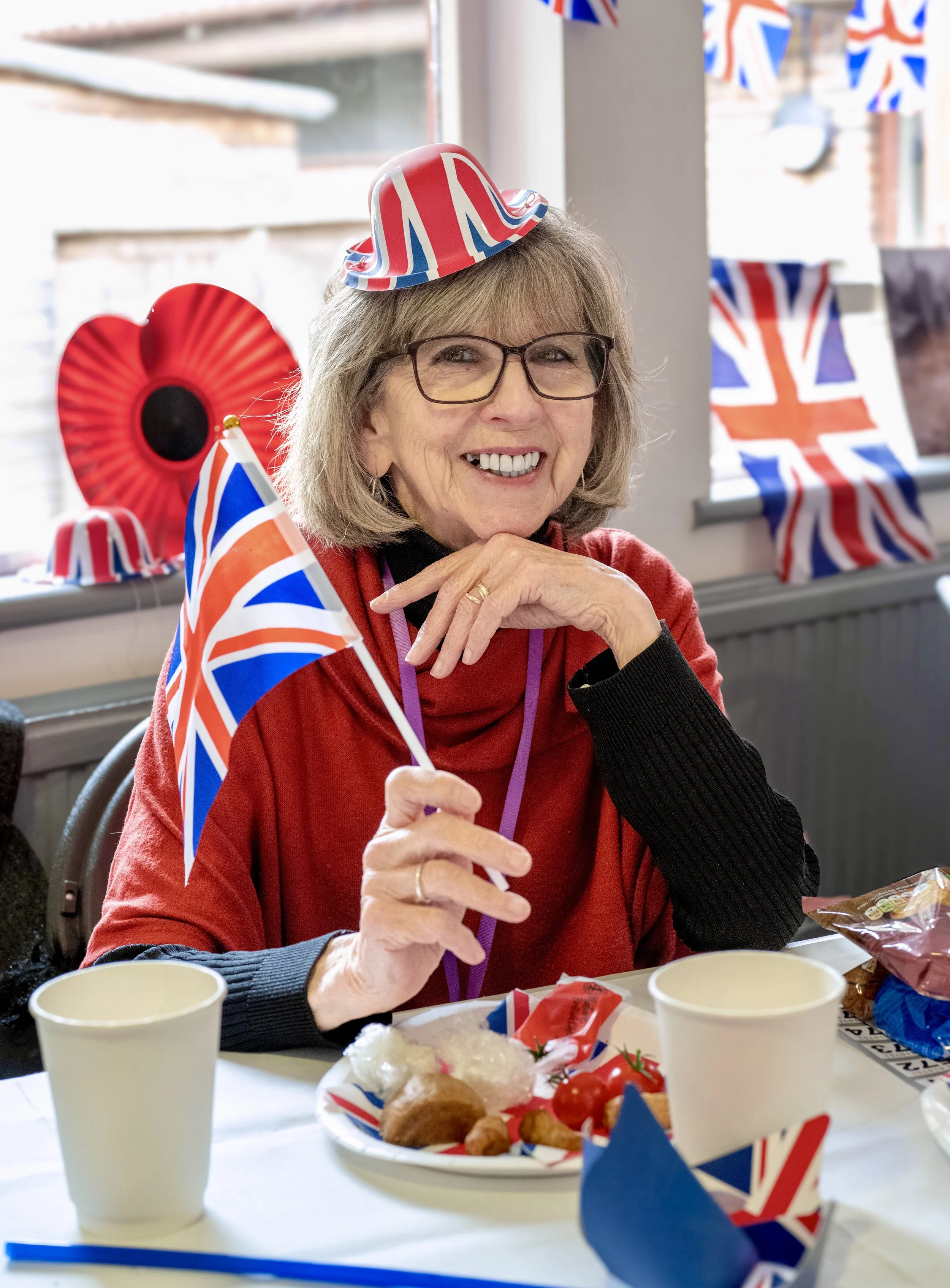 An elderly woman celebrating with Union Jack themed decorations, including a small British flag, at a party. She is smiling, wearing glasses, a red sweater, and a small Union Jack hat, sitting at a table with food, cups, and more Union Jack decoratio