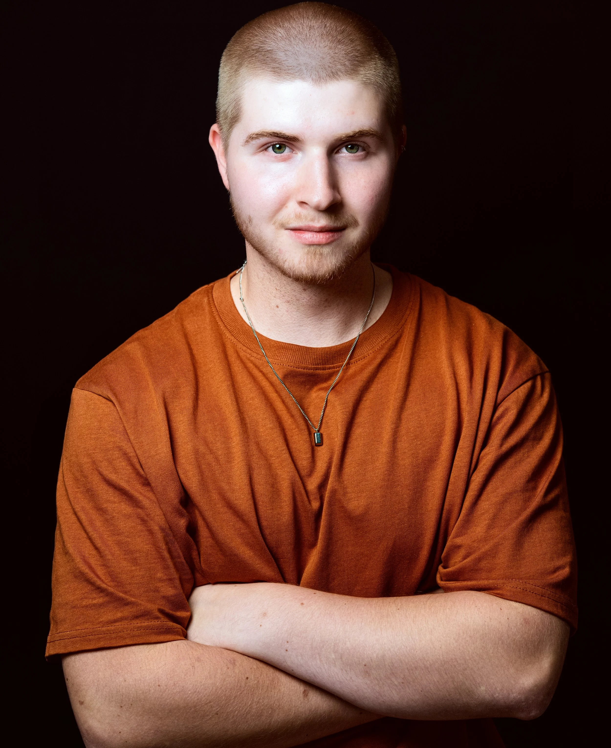 Young man with short blond hair and a light beard, wearing an orange T-shirt and a necklace, standing with arms crossed in front of a black background.