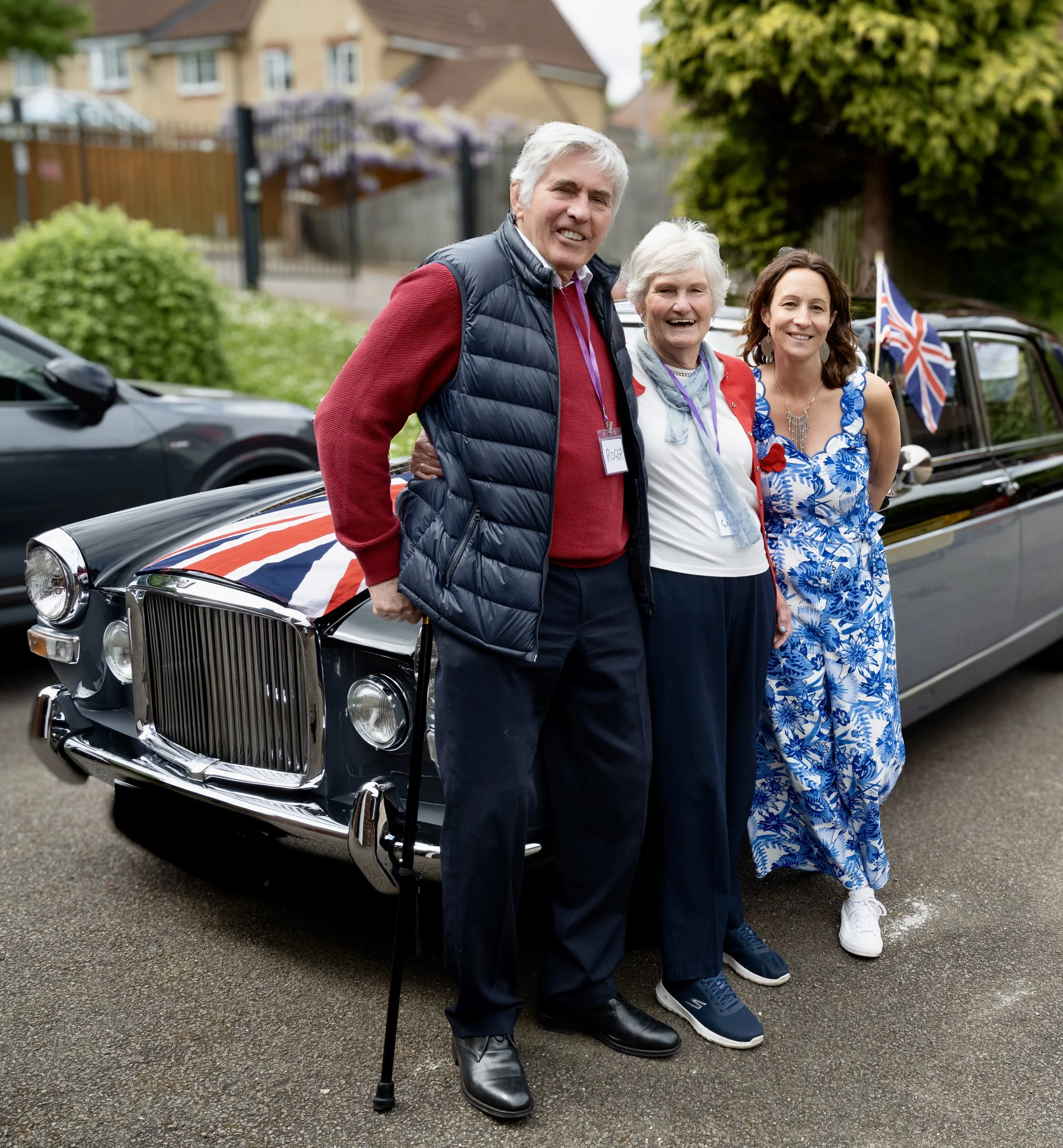 Three people standing in front of a vintage black car decorated with British flags, smiling, during a patriotic celebration outdoors.
