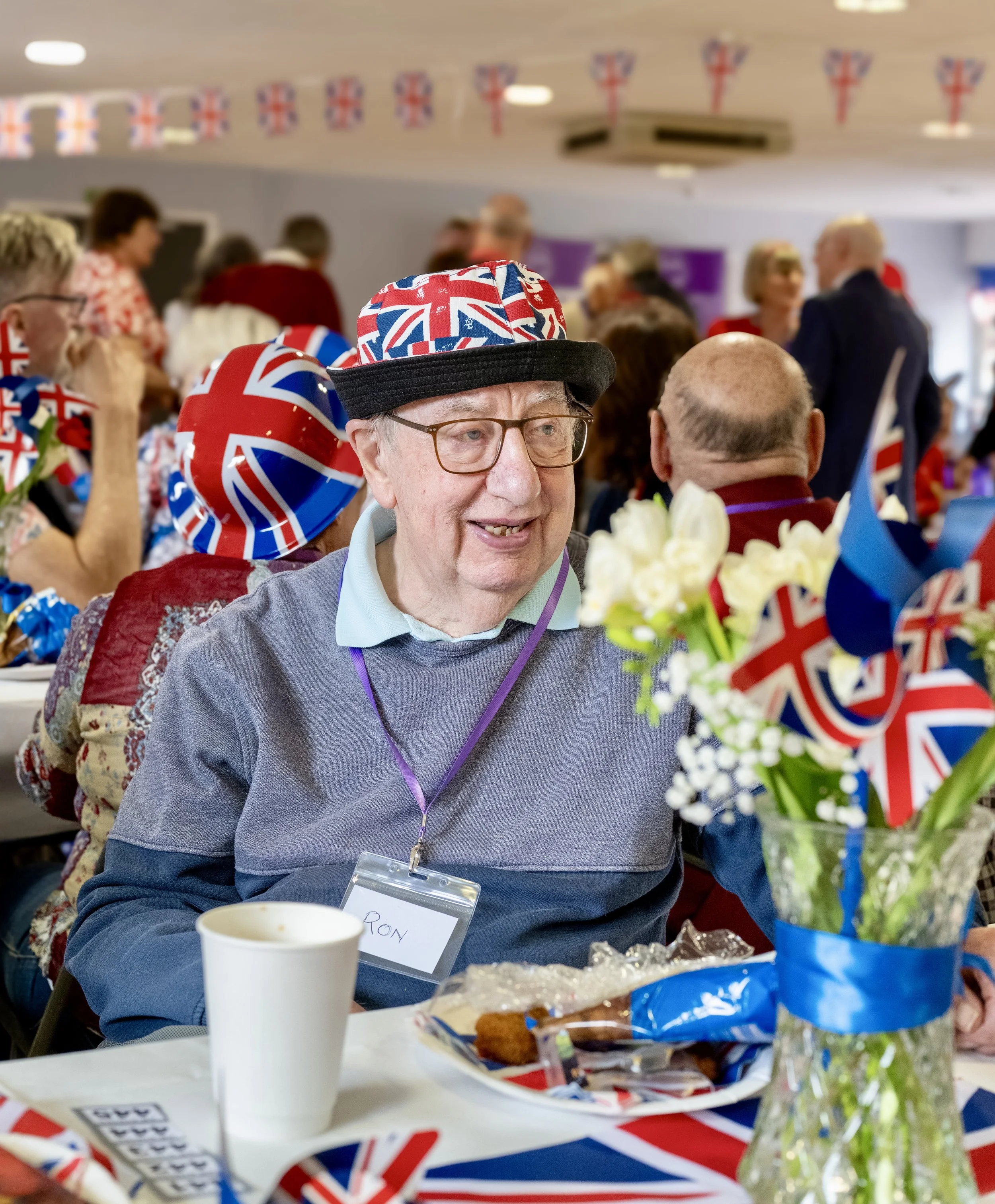 An elderly man wearing glasses, a hat with the Union Jack design, and a gray sweater, smiling during a celebration with Union Jack themed decorations, flags, and people in the background.
