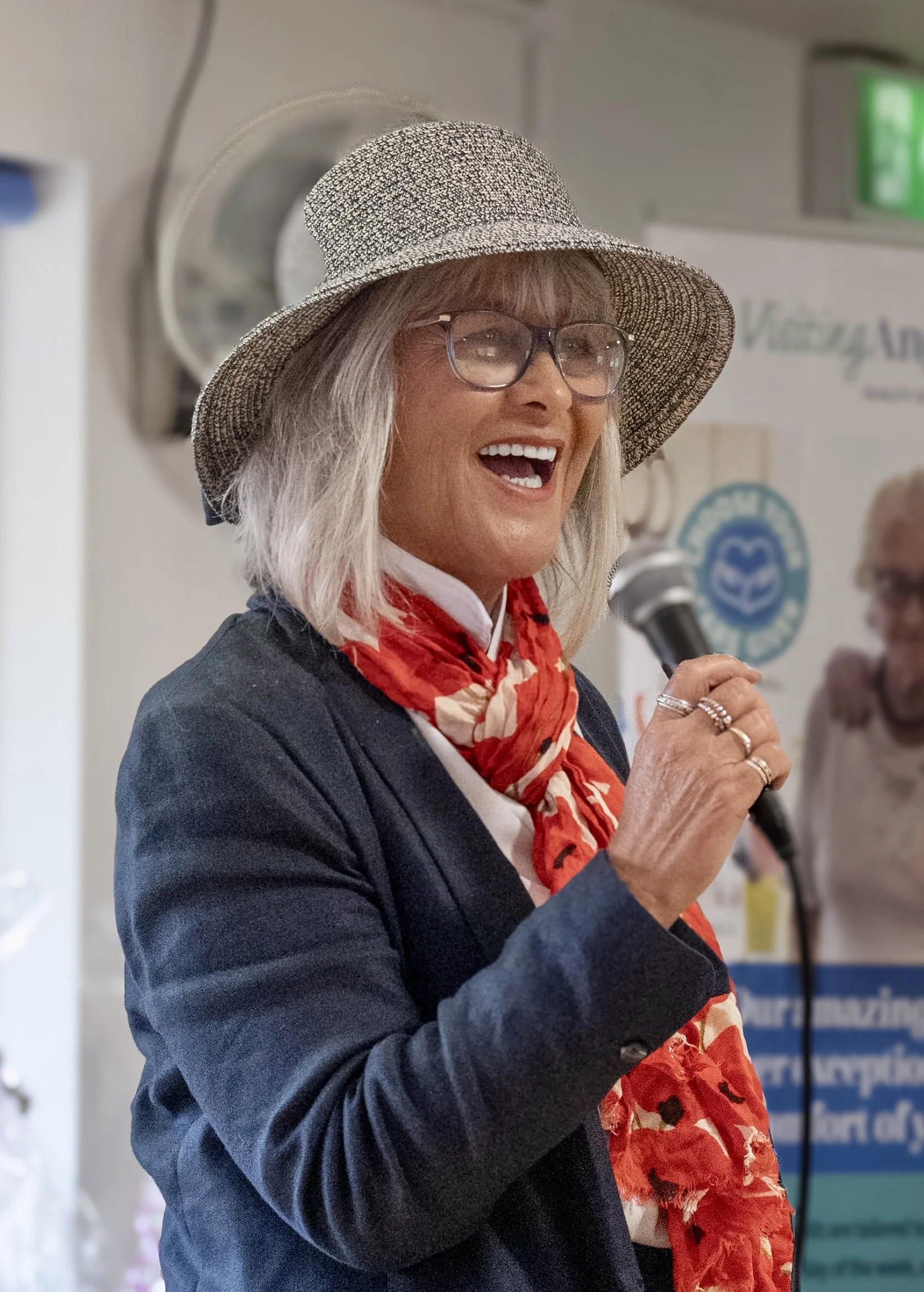 A smiling older woman wearing glasses, a wide-brimmed hat, a dark blazer, and a red scarf, holding a microphone and speaking at an indoor event.