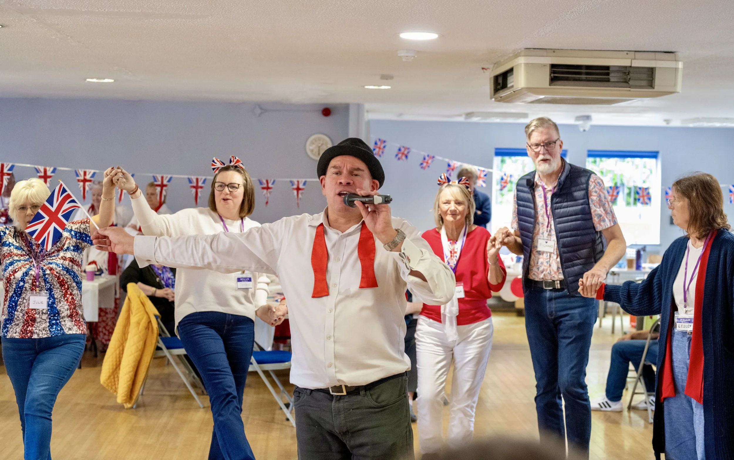 Man singing into a microphone at a celebration with Union Jack flags and people dancing.