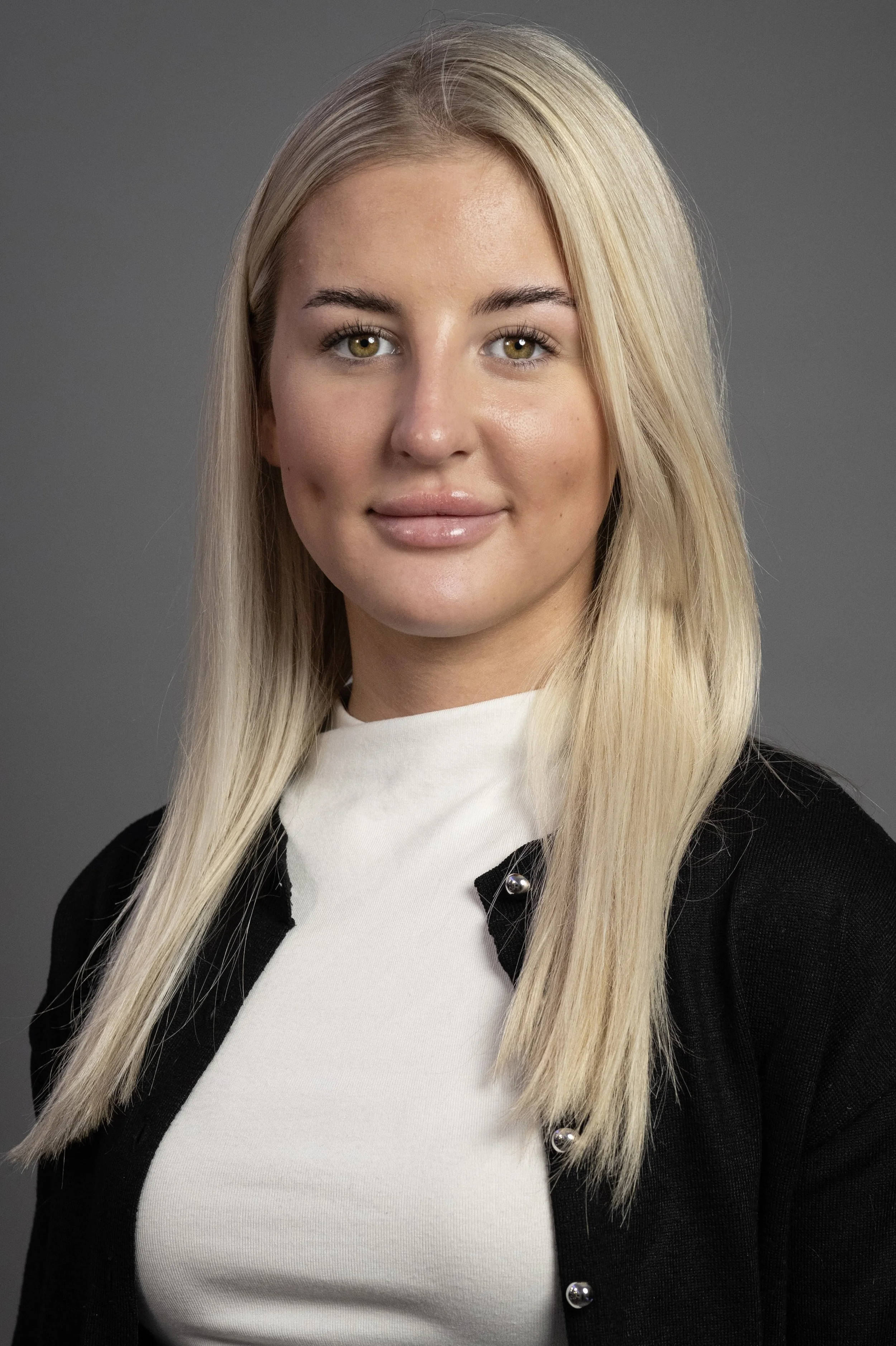 A young woman with long blonde hair, light skin, and hazel eyes, wearing a white top and a black cardigan, posing against a gray background. Corporate Headshot