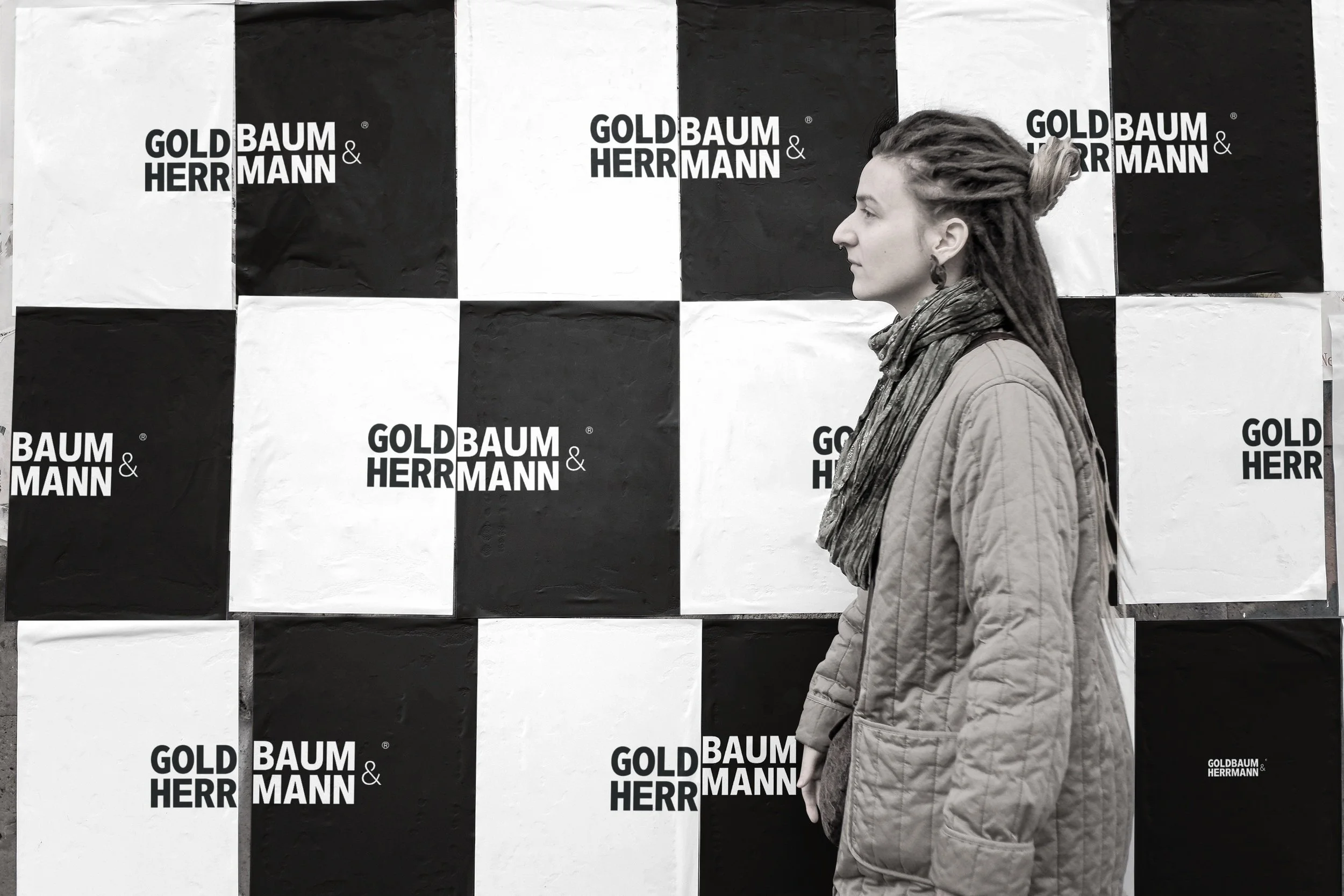A woman with dreadlocks in profile standing in front of a black and white checkerboard backdrop with the logos 'GOLDBAUM & HERRMANN'.