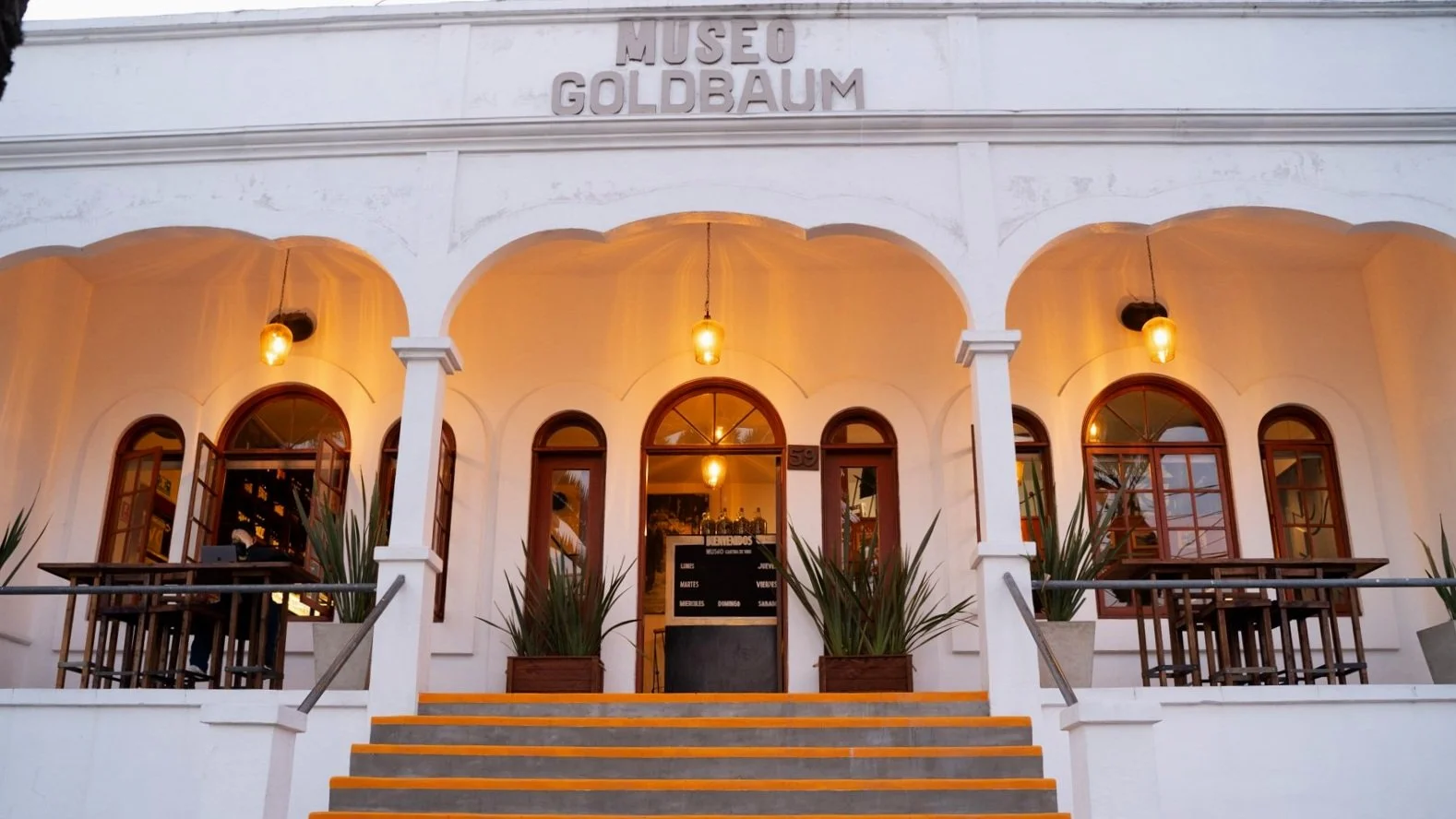 Front view of Museo Goldbaum, with lights hanging from the ceiling, arched windows, potted plants on either side of the entrance, and orange steps leading up to the door.