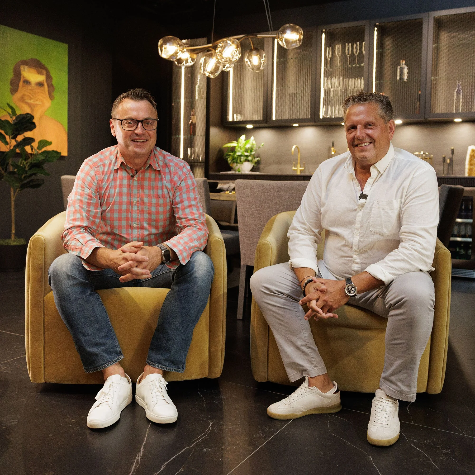 Two men sitting in yellow armchairs in a modern kitchen or lounge area, smiling at the camera.