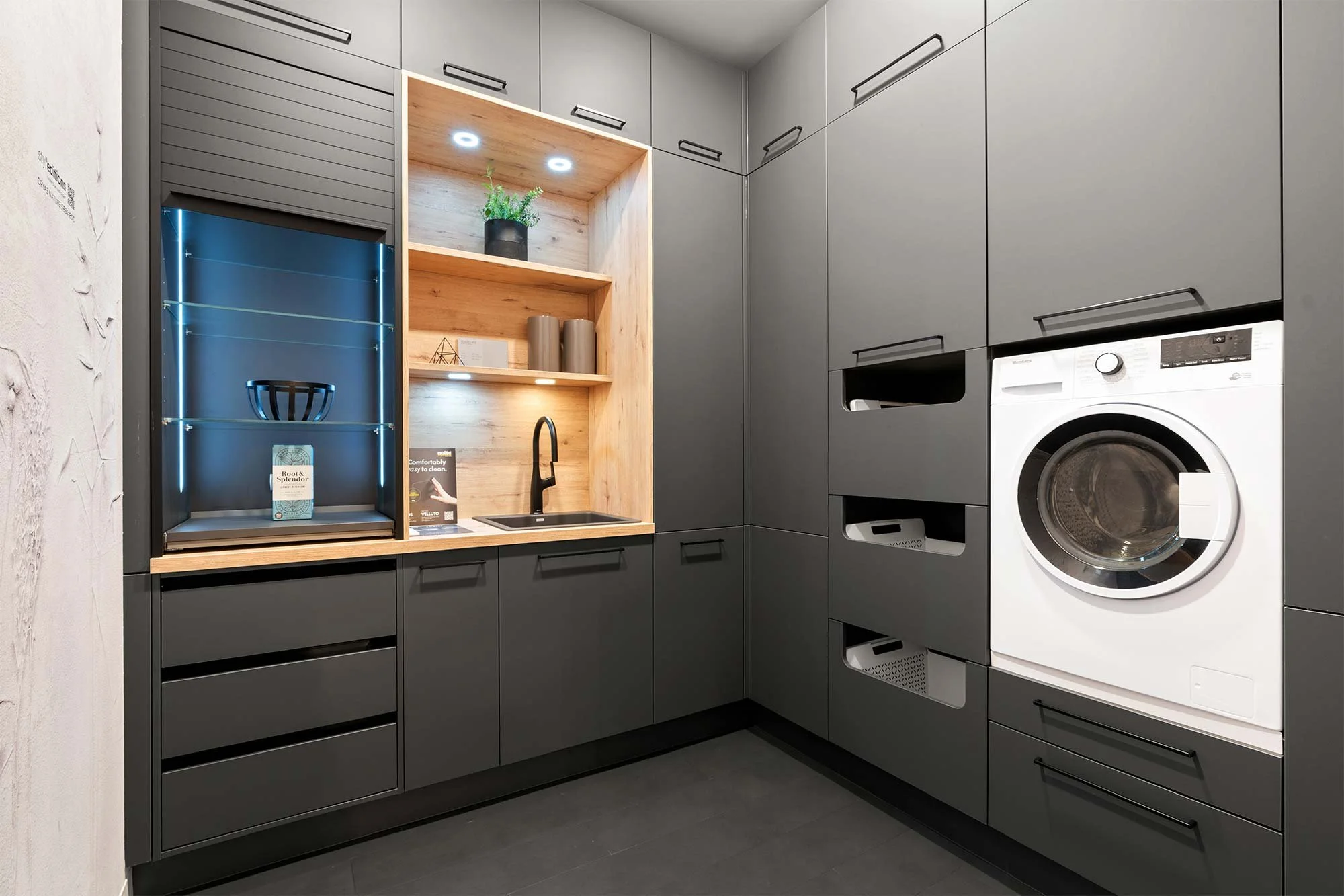 Modern kitchen with white walls, gray island, and open shelving, woman cooking at the island, large window with sunlight, dining table with bowl and decorative items, plants and artwork in the space.