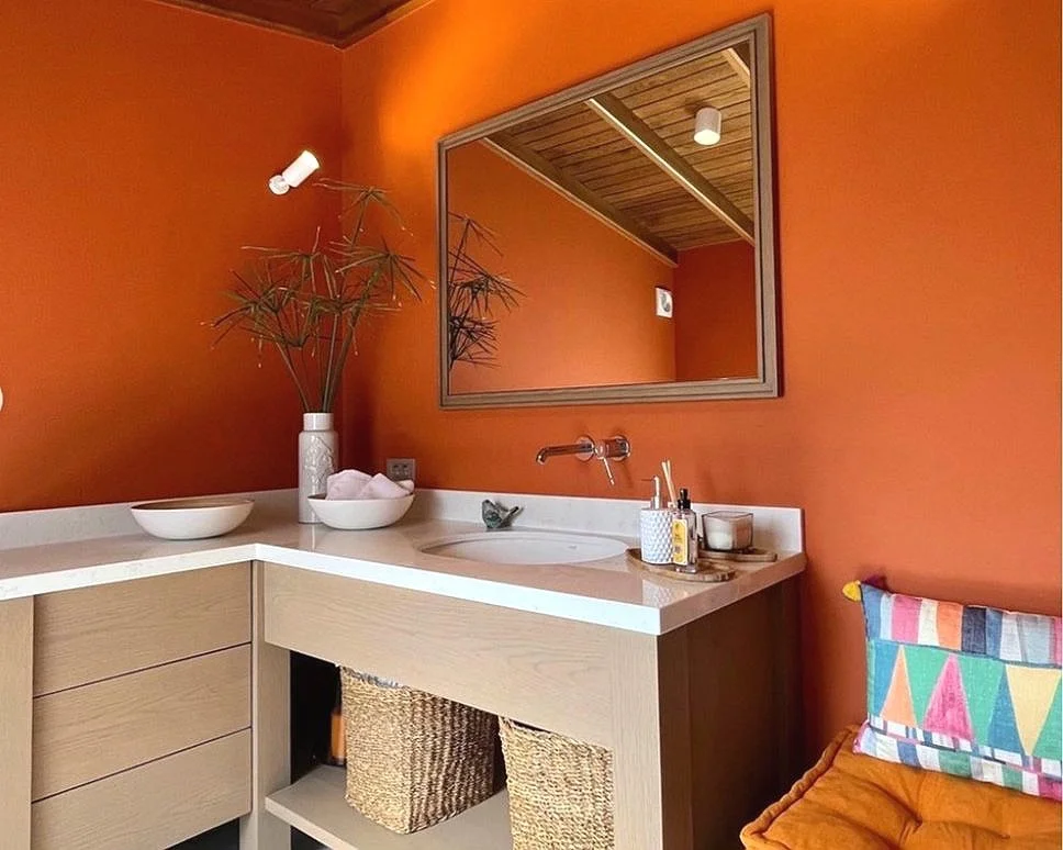 A vibrant bathroom with wooden ceiling, oak cabinets, a white countertop, a signature orange wall paint and neutral, minimalist accessories.