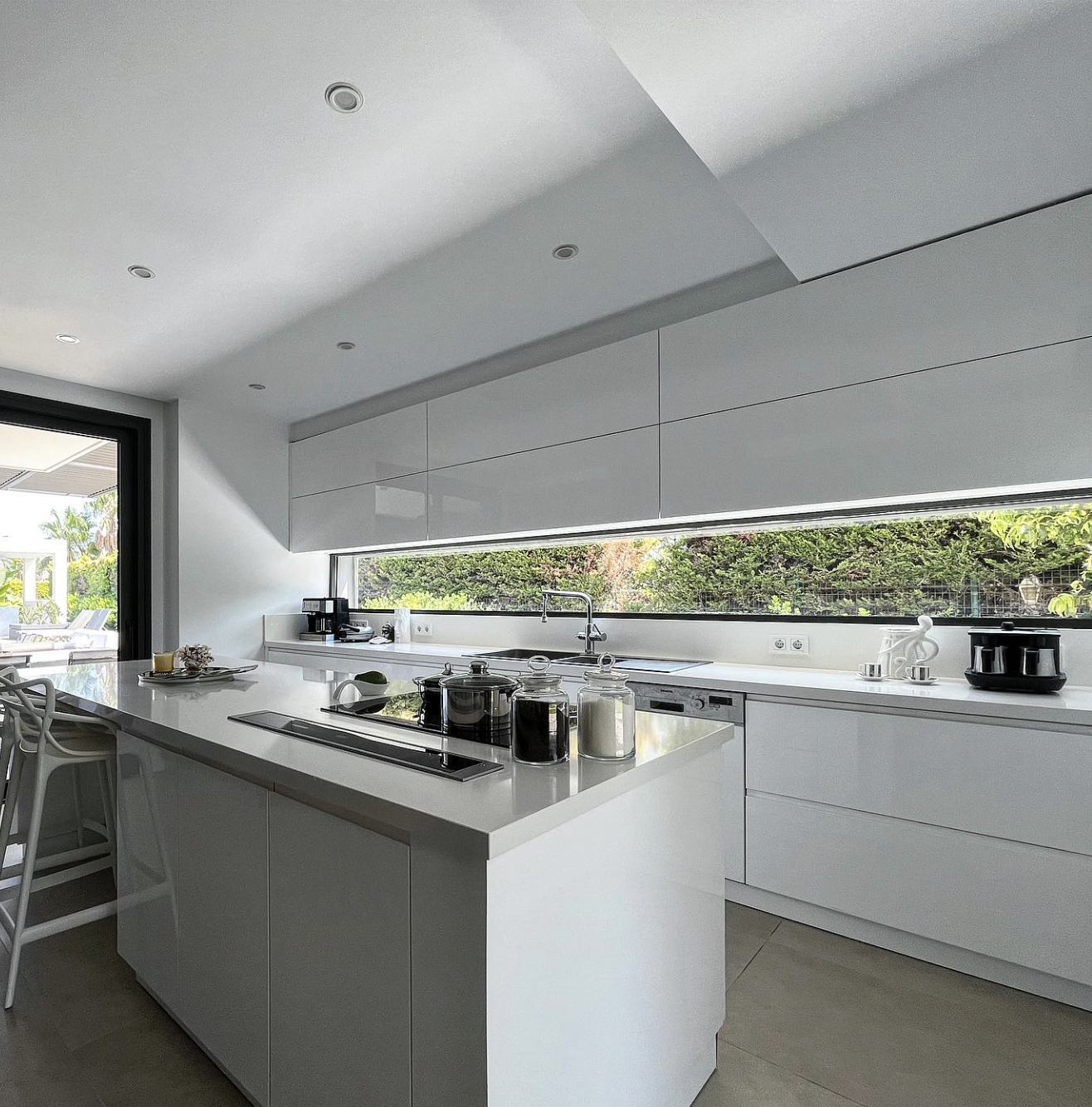 Modern minimalist kitchen with central island and white cabinets. Large windows create a bright and airy ambiance. The reflective surfaces of the white cabinets and quartz countertop amplify the natural light.
