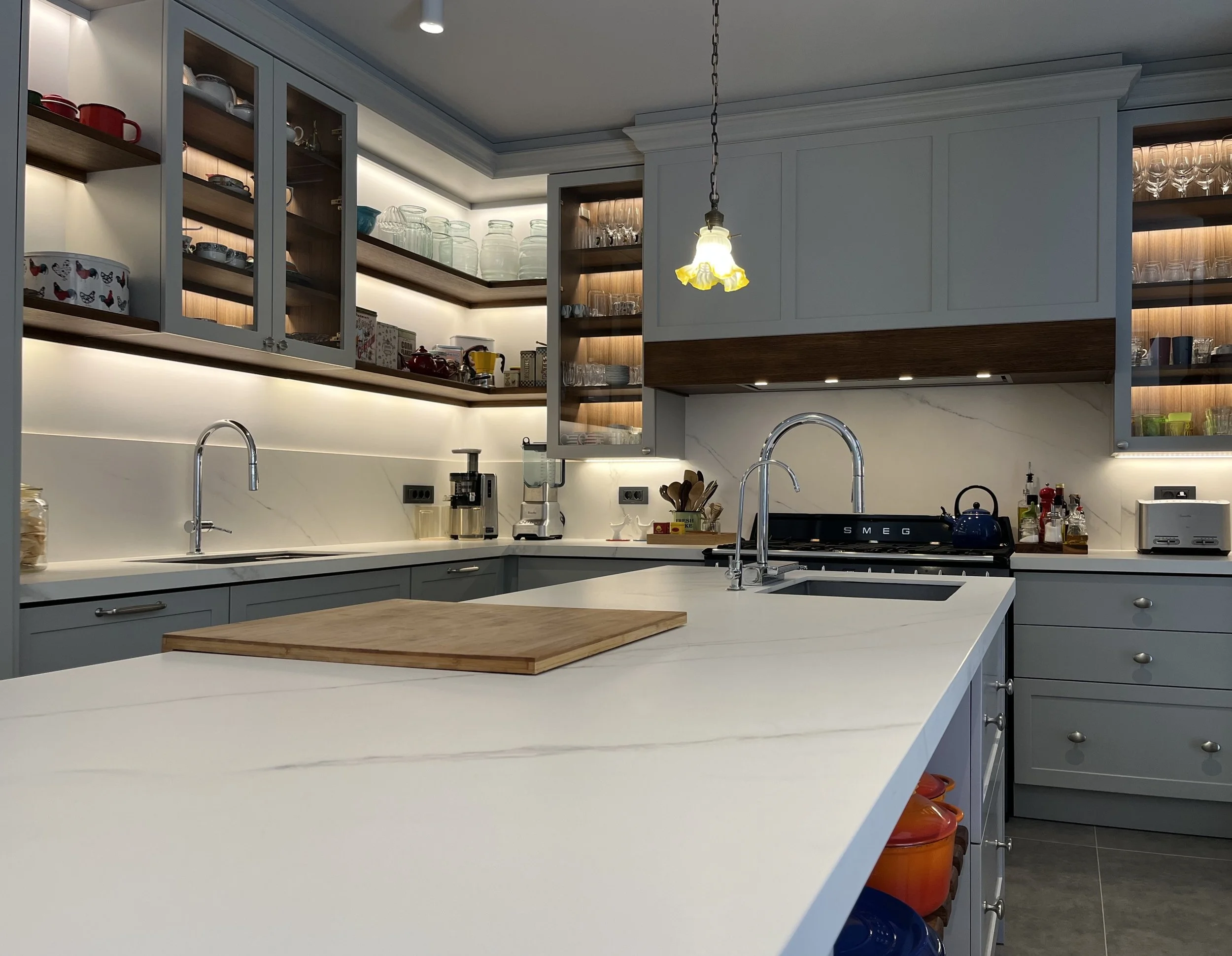 Eclectic modern kitchen featuring soft-grey lacquered HDF shaker cabinetry, warm wood accents, and a marble-veined Cosentino quartz backsplash - central large island detail. 