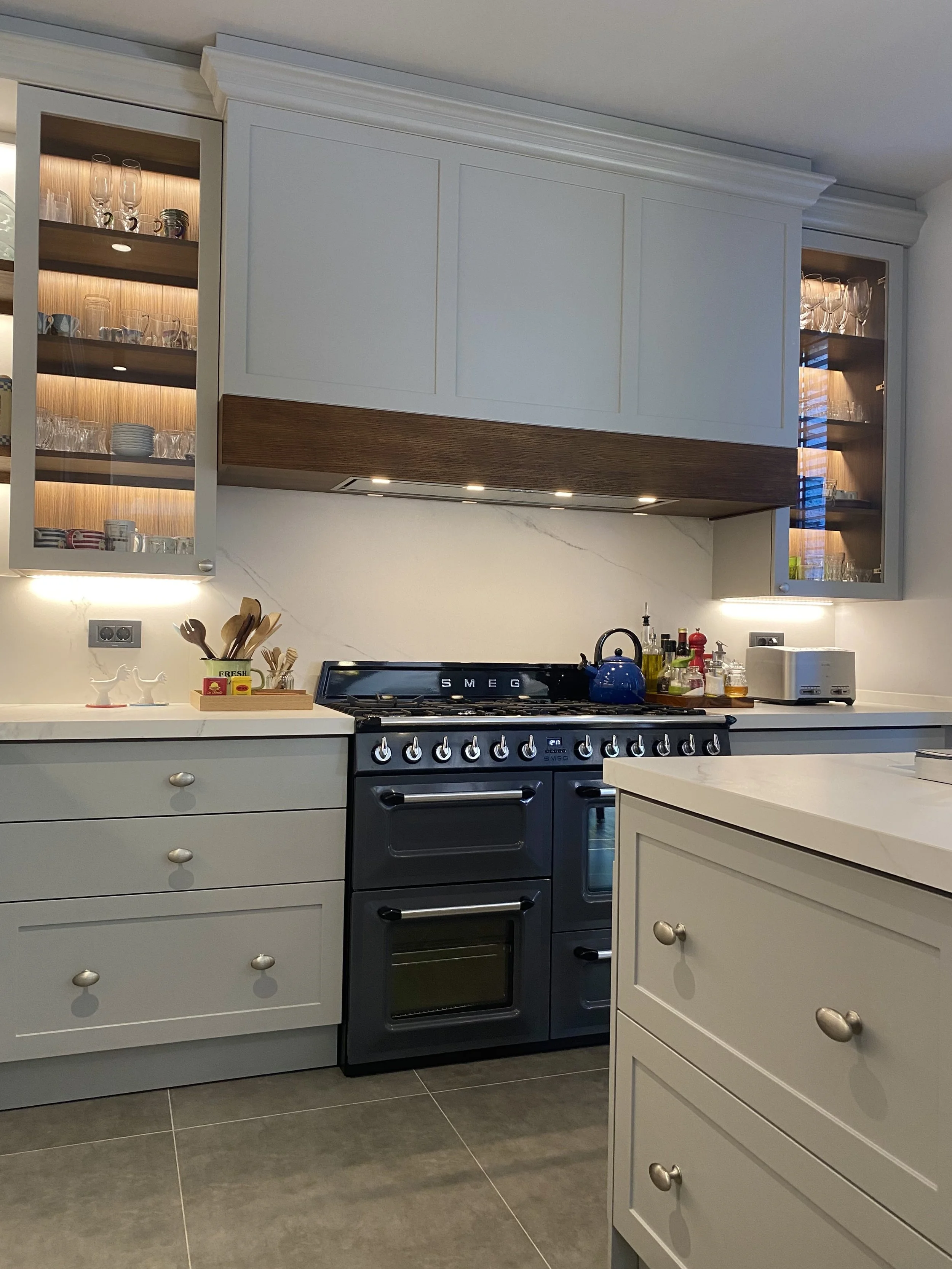 Eclectic modern kitchen featuring soft-grey lacquered HDF shaker cabinetry, warm wood accents, and a marble-veined Cosentino quartz backsplash, with integrated LED lighting. 