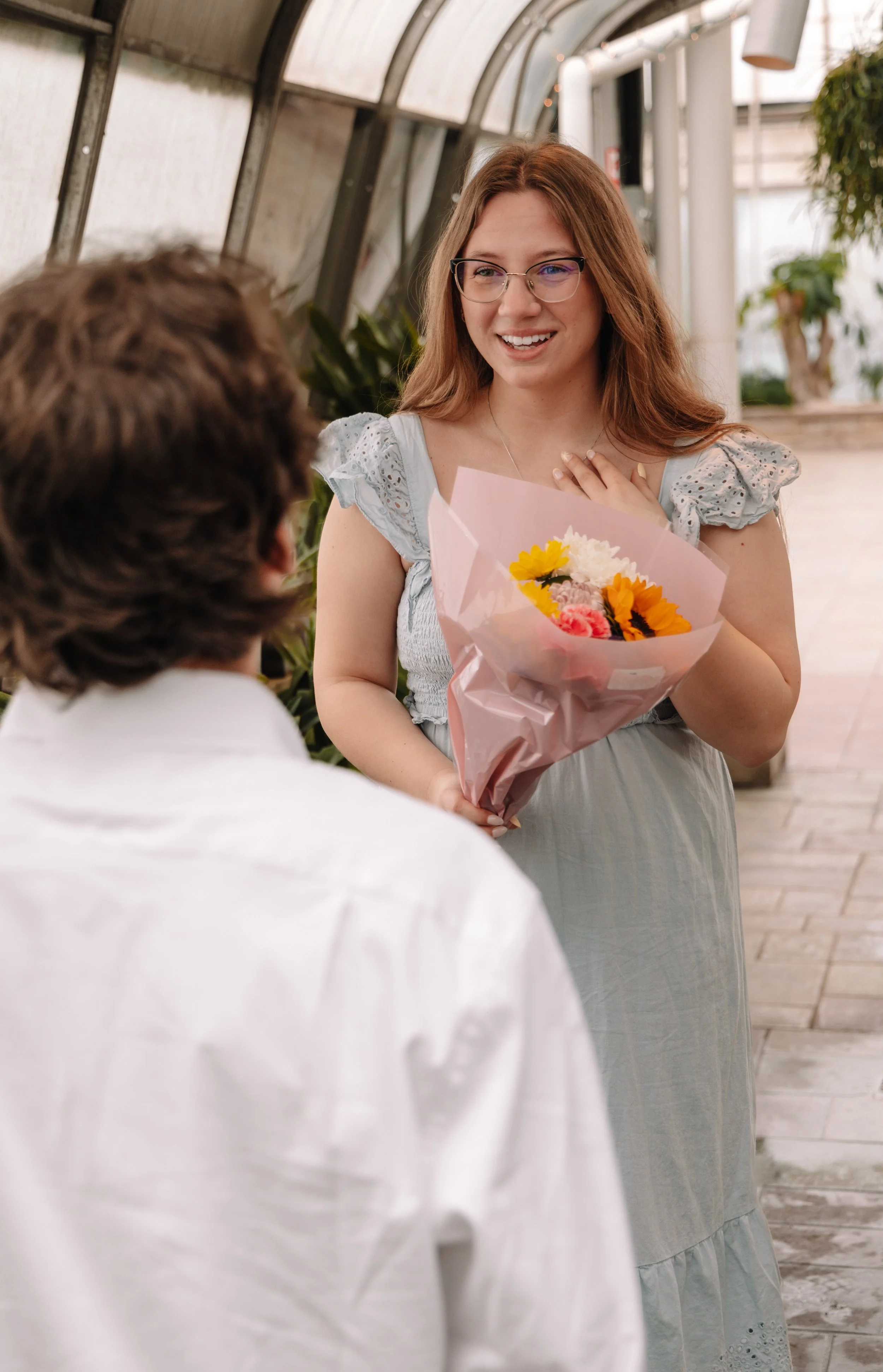 A woman with long red hair, glasses, and a light blue dress holding a bouquet of flowers and smiling at a man with brown hair in a white shirt. They are inside a greenhouse or botanical garden.