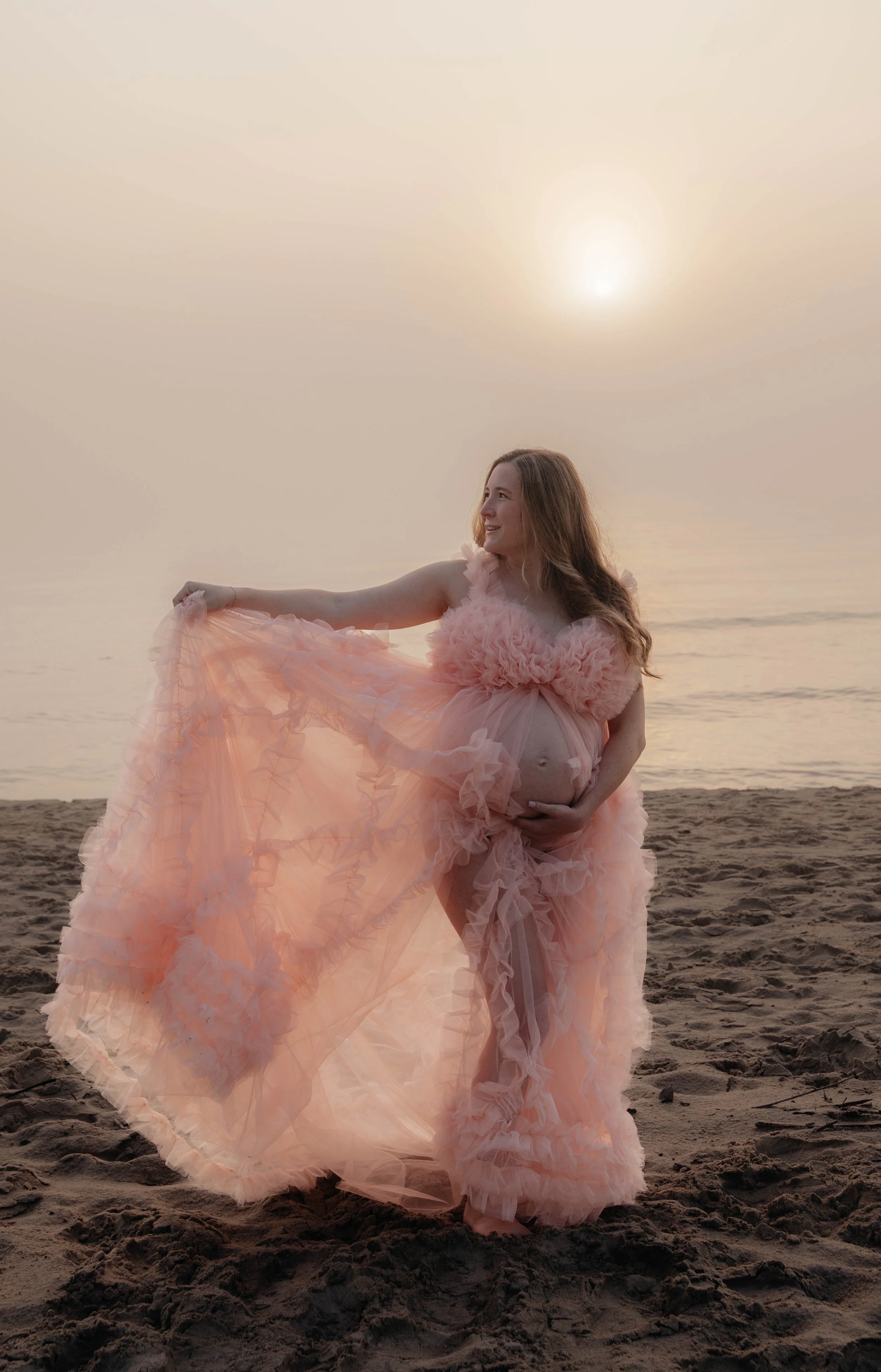 Pregnant woman in a peach, ruffled gown standing on a sandy beach at sunset.