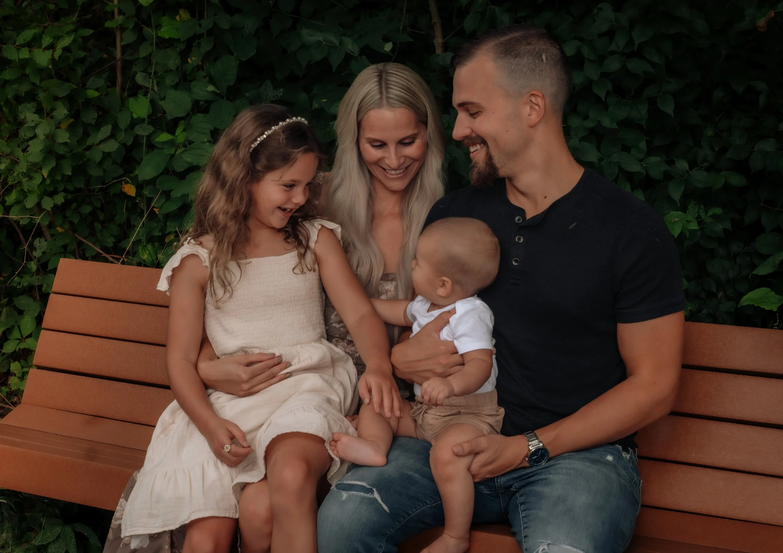 A smiling family sitting on a wooden park bench with green leafy bushes in the background, enjoying a moment together. The family includes a woman, a man, a young girl, and a baby.
