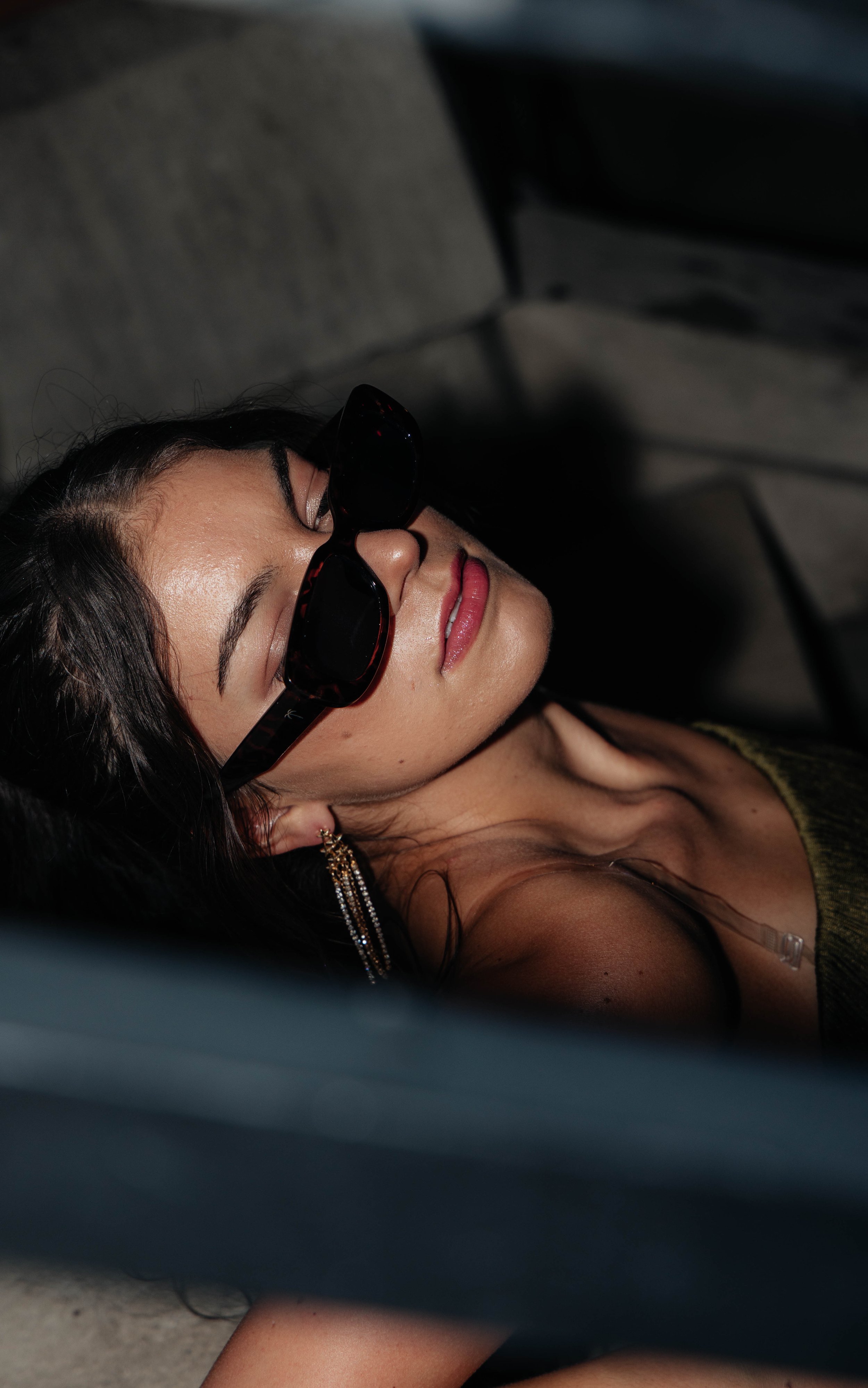 A woman with dark hair and makeup, wearing sunglasses, braces, and gold earrings, lying on the ground with a neutral expression.