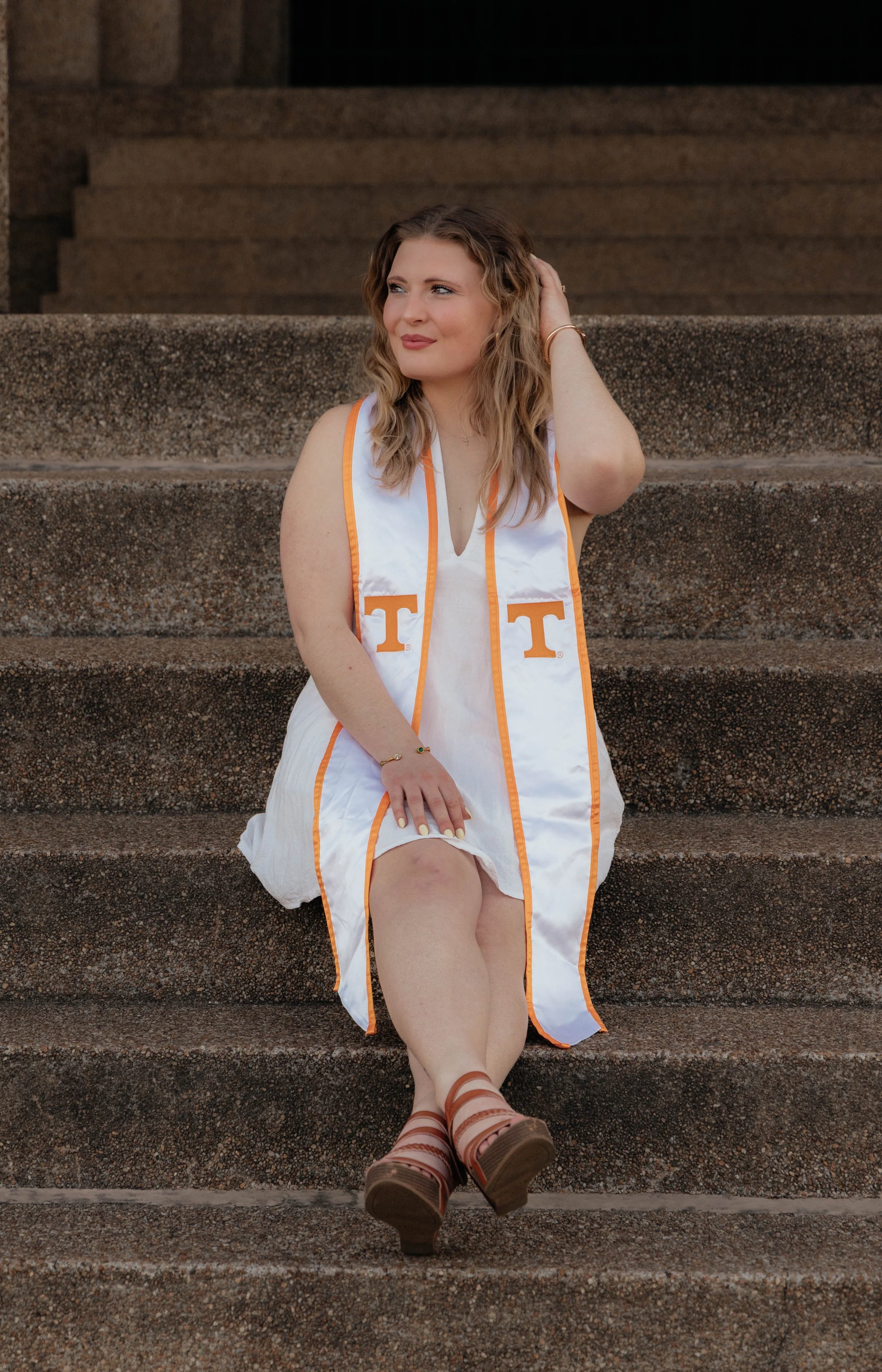A woman sitting on outdoor stone steps wearing a white dress and a university graduation stole with the letter T.