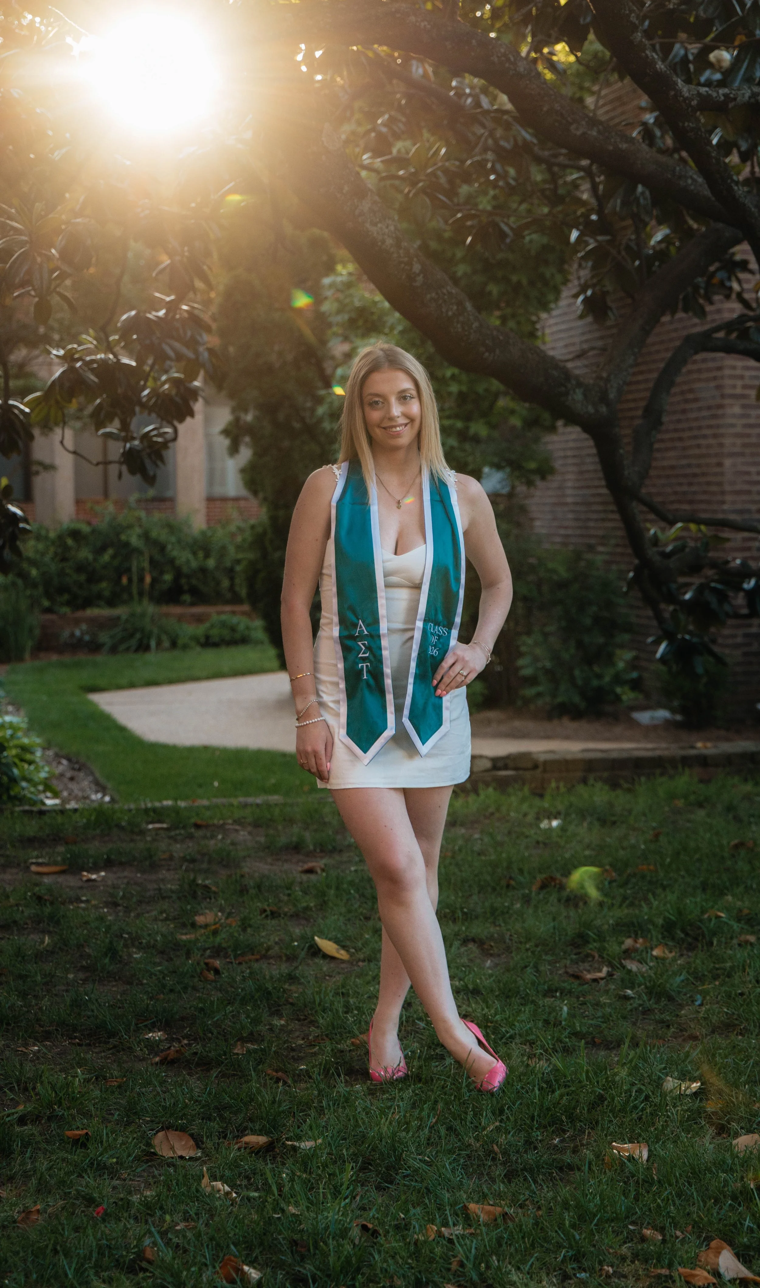 A young woman wearing a white dress and pink heels, smiling and standing outdoors on a grassy area with trees and a brick building in the background. She has a teal graduation stole with Greek letters and the year 2026.