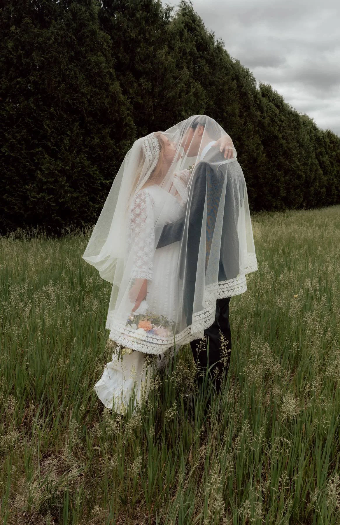A bride and groom sharing a kiss in a field, with the bride's veil covering both of their faces.
